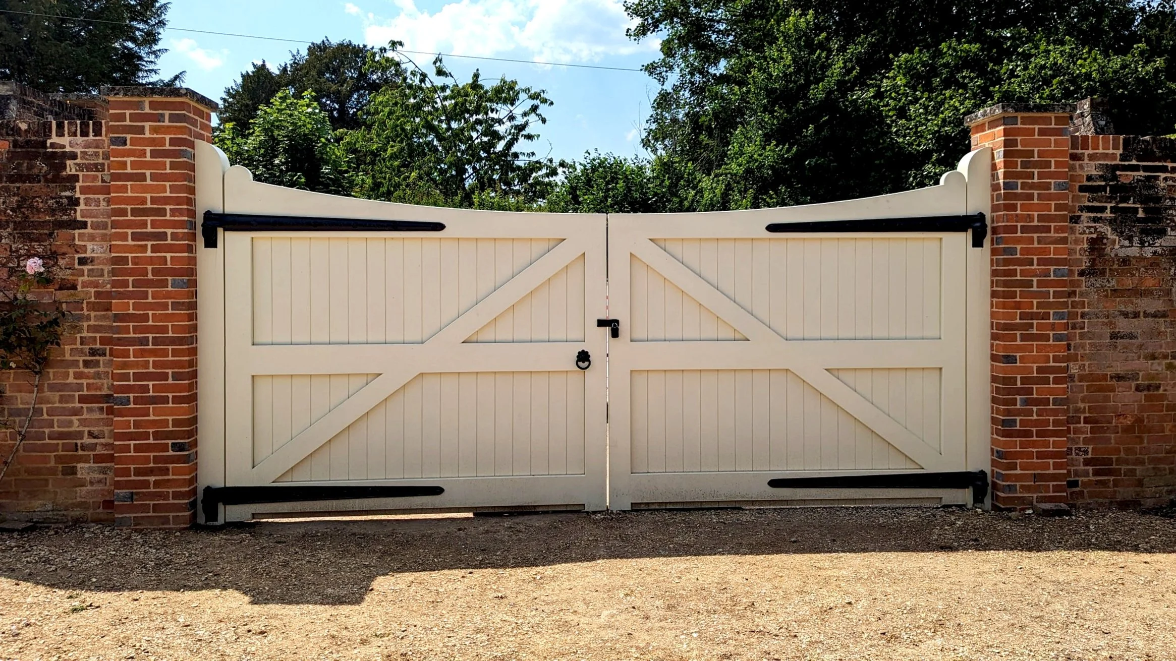 White double gate with black hinges and latch, flanked by brick pillars, with trees and blue sky in the background.