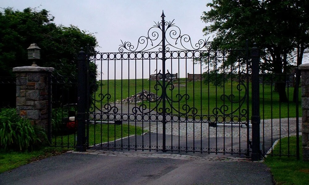Black wrought iron gate with decorative scrollwork, flanked by stone pillars, opening to a garden with a cobblestone path, green grass, trees, and a bench in the background.