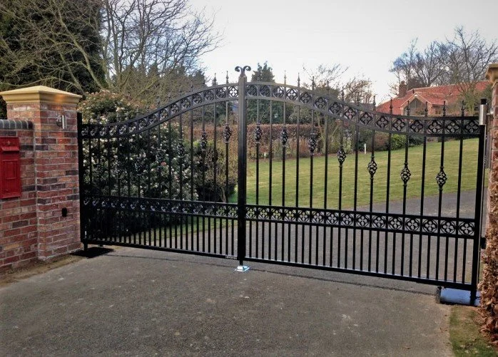 Black wrought iron gate with decorative elements, set between brick pillars, opening onto a paved driveway and a grassy lawn with trees and houses in the background.