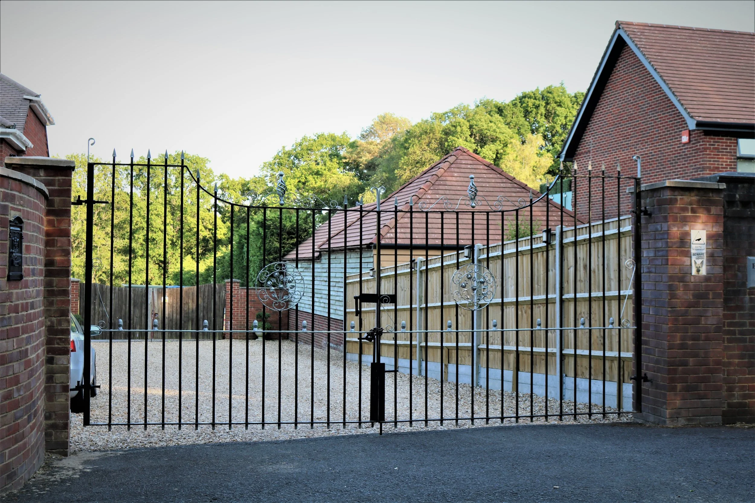 Residential driveway with a black metal gate, brick wall pillars, houses with brick walls and tiled roofs, and green trees in the background.