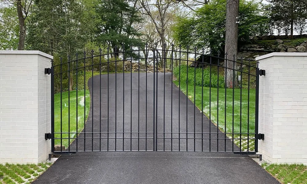 Black metal driveway gate between white brick pillars, leading to a paved driveway surrounded by green grass and trees.