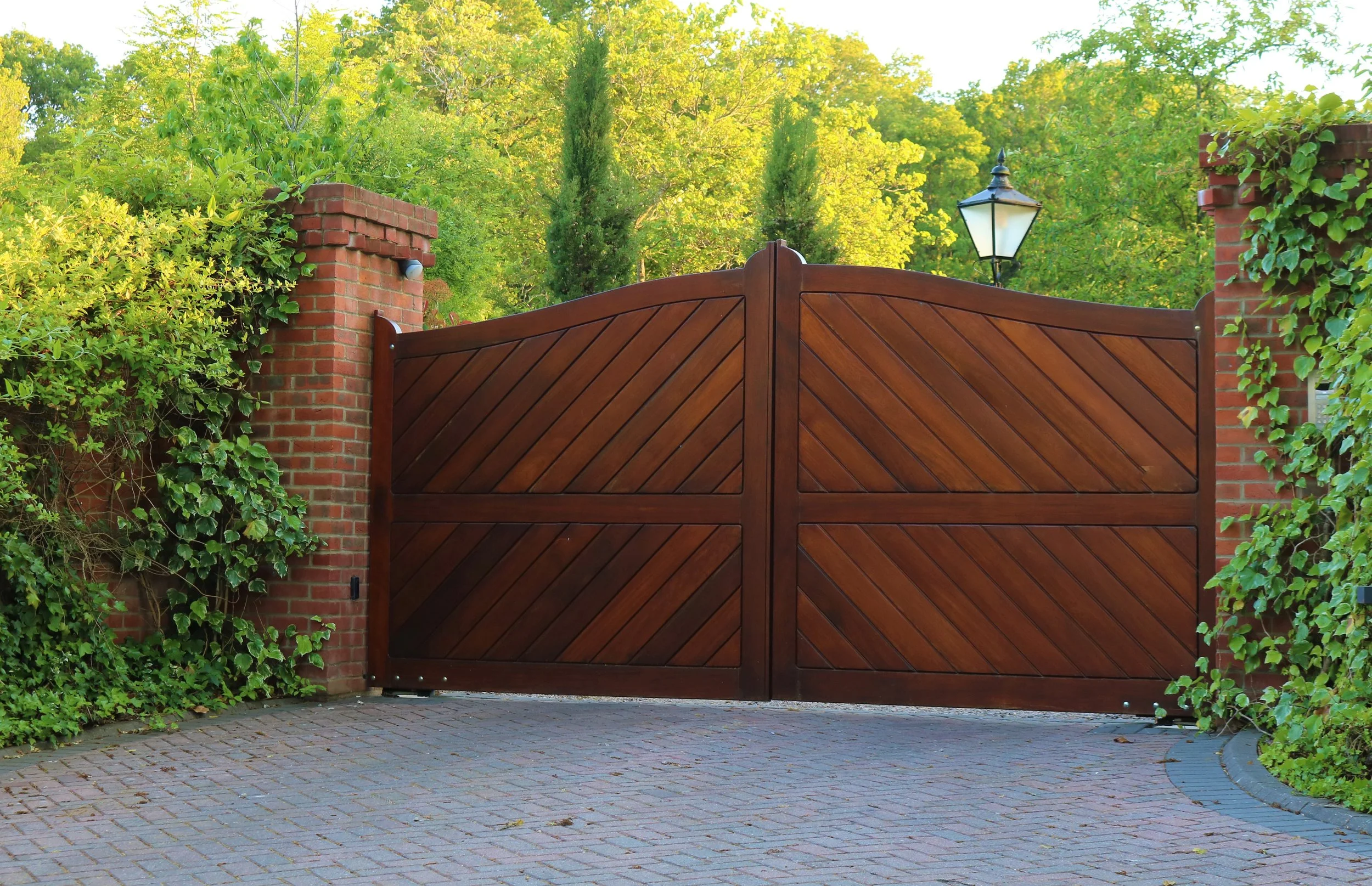 A wooden double gate with diagonal planks, flanked by brick columns covered in green ivy, with a black lantern-style light mounted on the right column, set against a backdrop of lush green trees and shrubs.