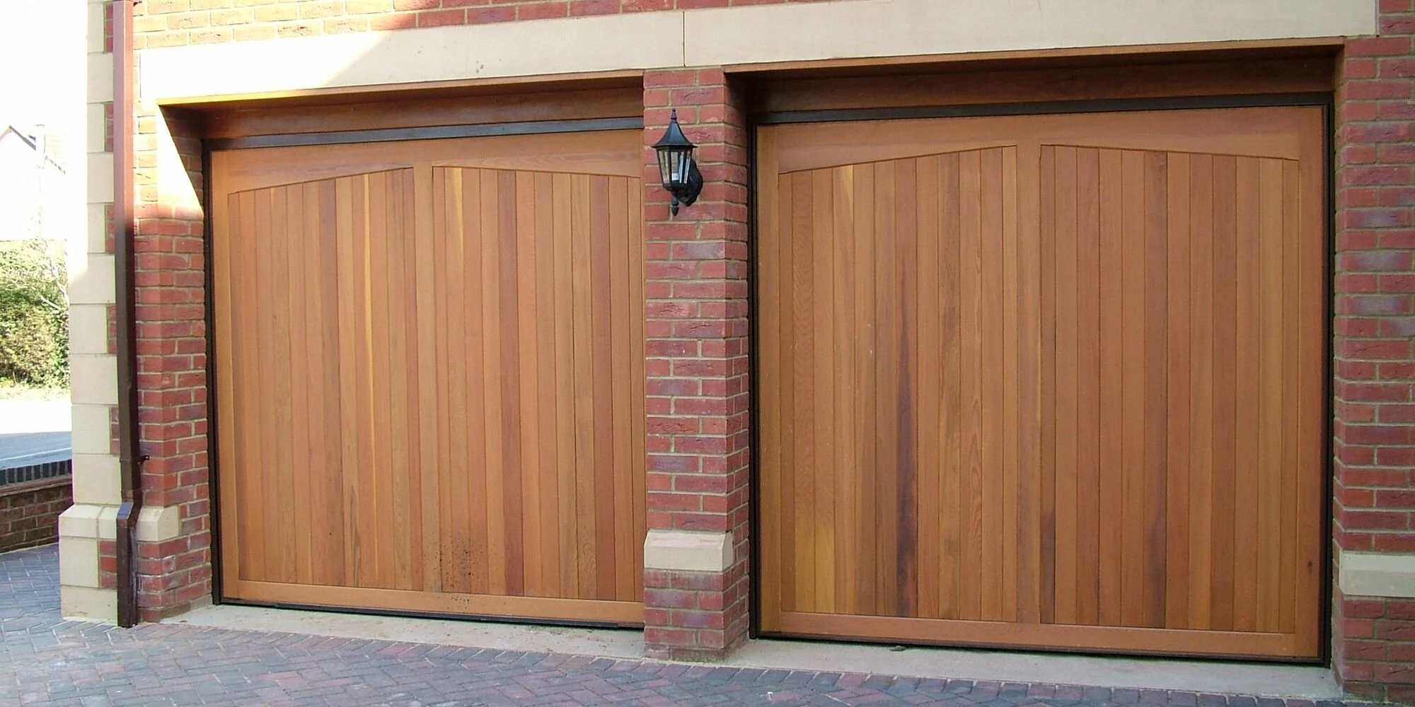 Two wooden garage doors with modern black trim, surrounded by red brick walls, with a black wall-mounted outdoor lantern beside the left door.