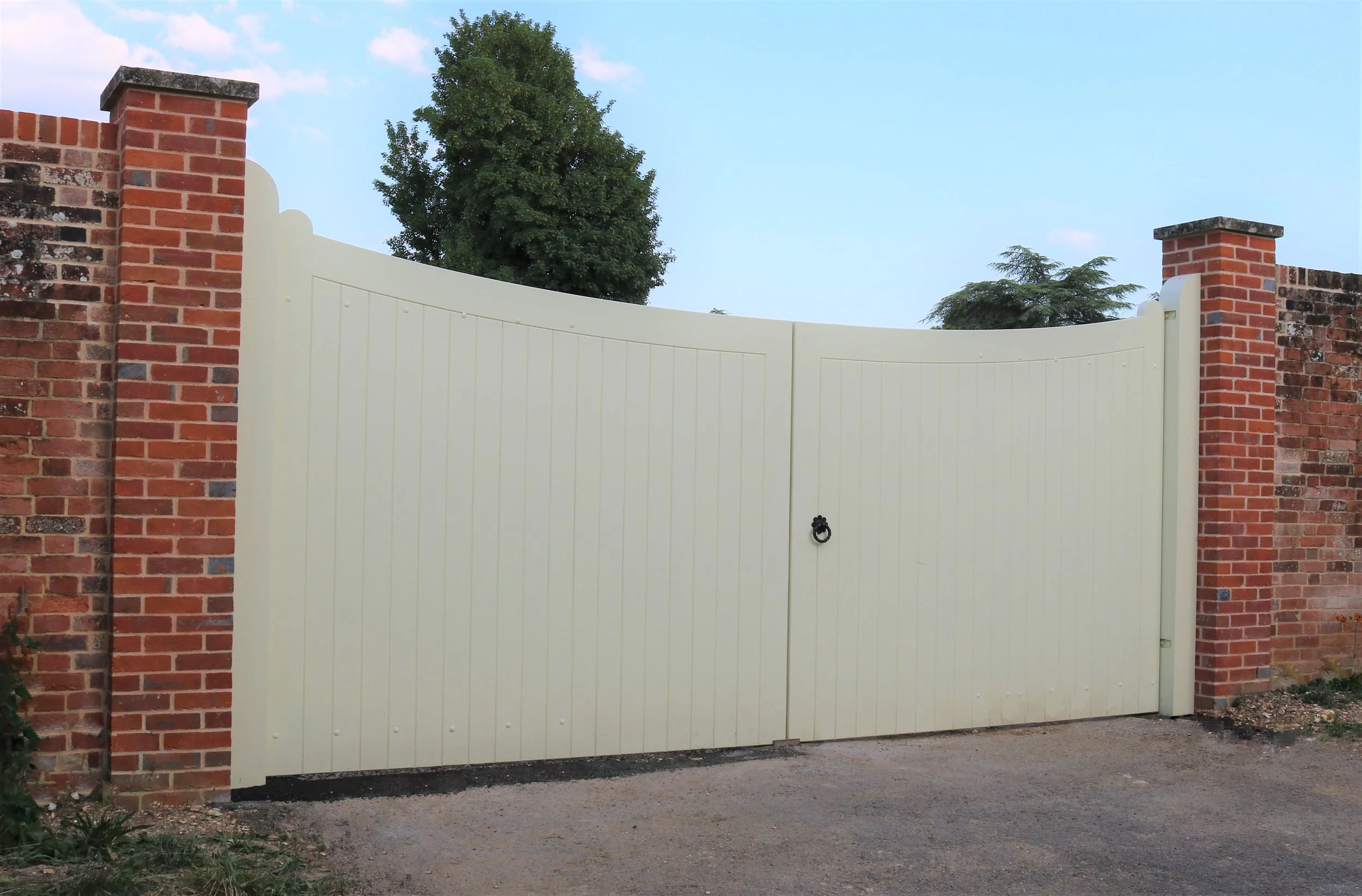 A white double gate in front of a brick wall with brick columns on each side. Tall trees are visible in the background under a clear blue sky.