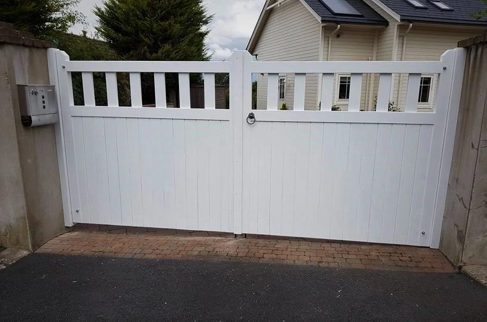 White double gate with a latch and a mailbox on the left side, installed between concrete walls on a paved surface, leading into a residential driveway.