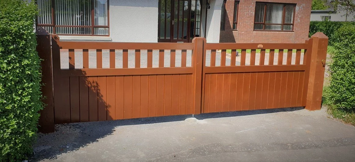 Brown wooden double gate in front of a house with a brick and stucco exterior and large windows, surrounded by greenery.