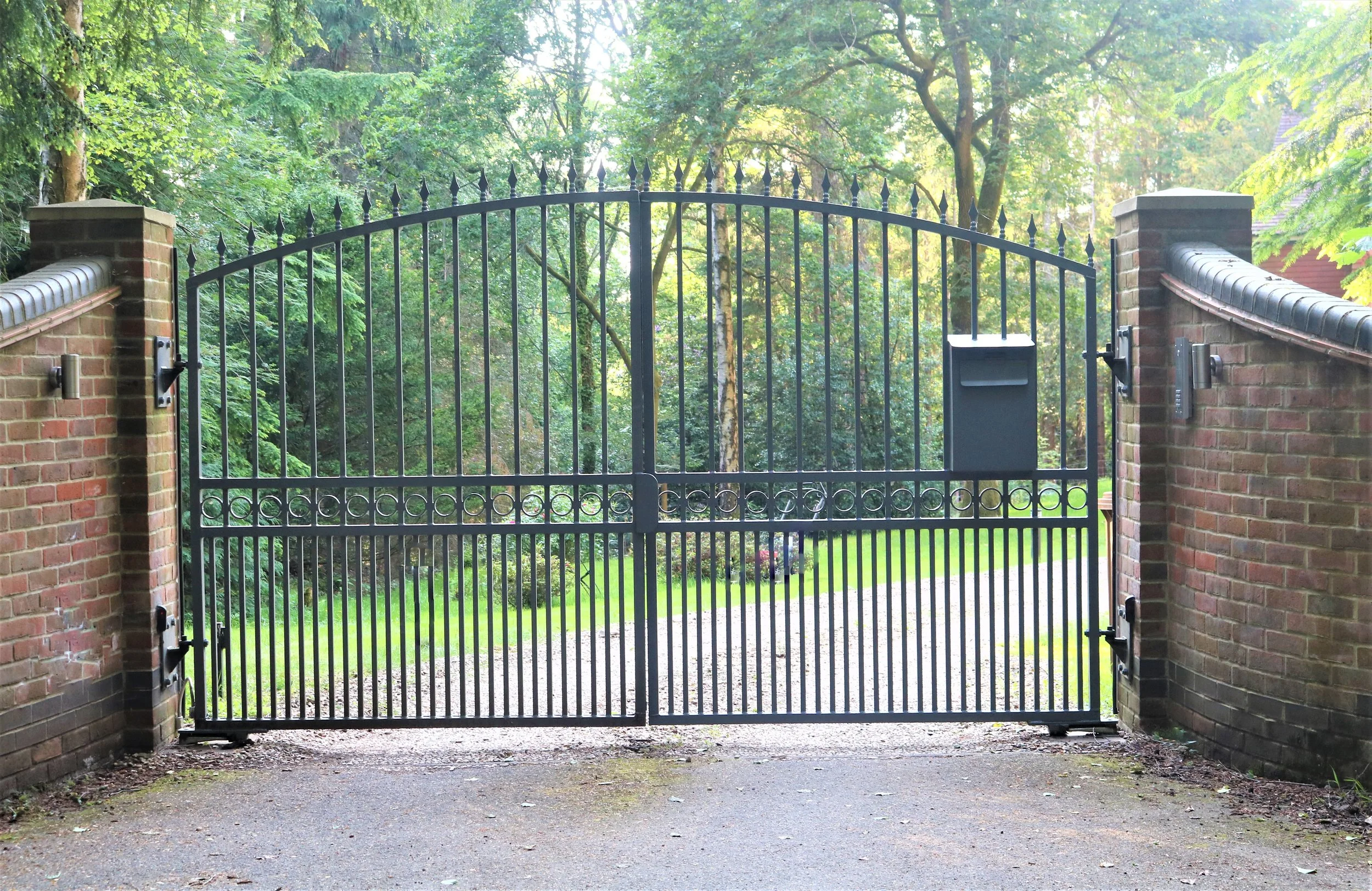 A black metal gate with pointed tops, flanked by brick pillars, opens to a green yard with trees and grass in the background.