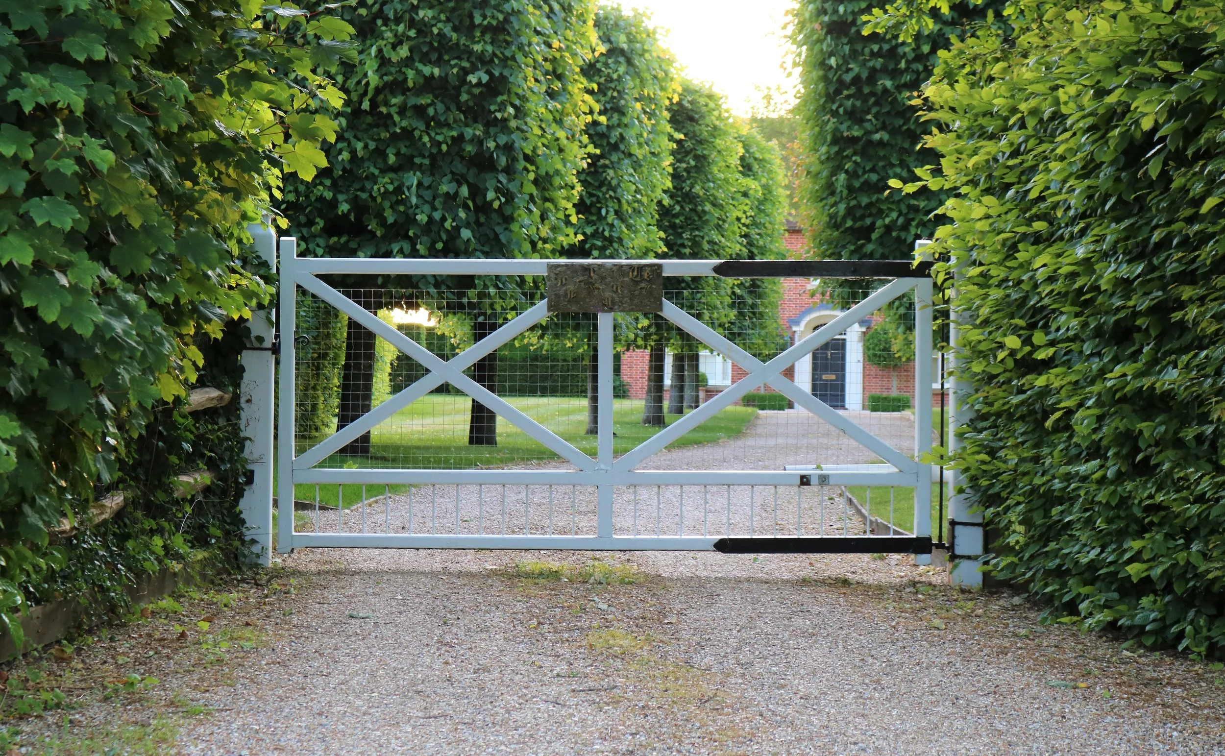 White gate with a wooden plaque, surrounded by green hedges, leading to a residential street with red brick house and green trees in the background.