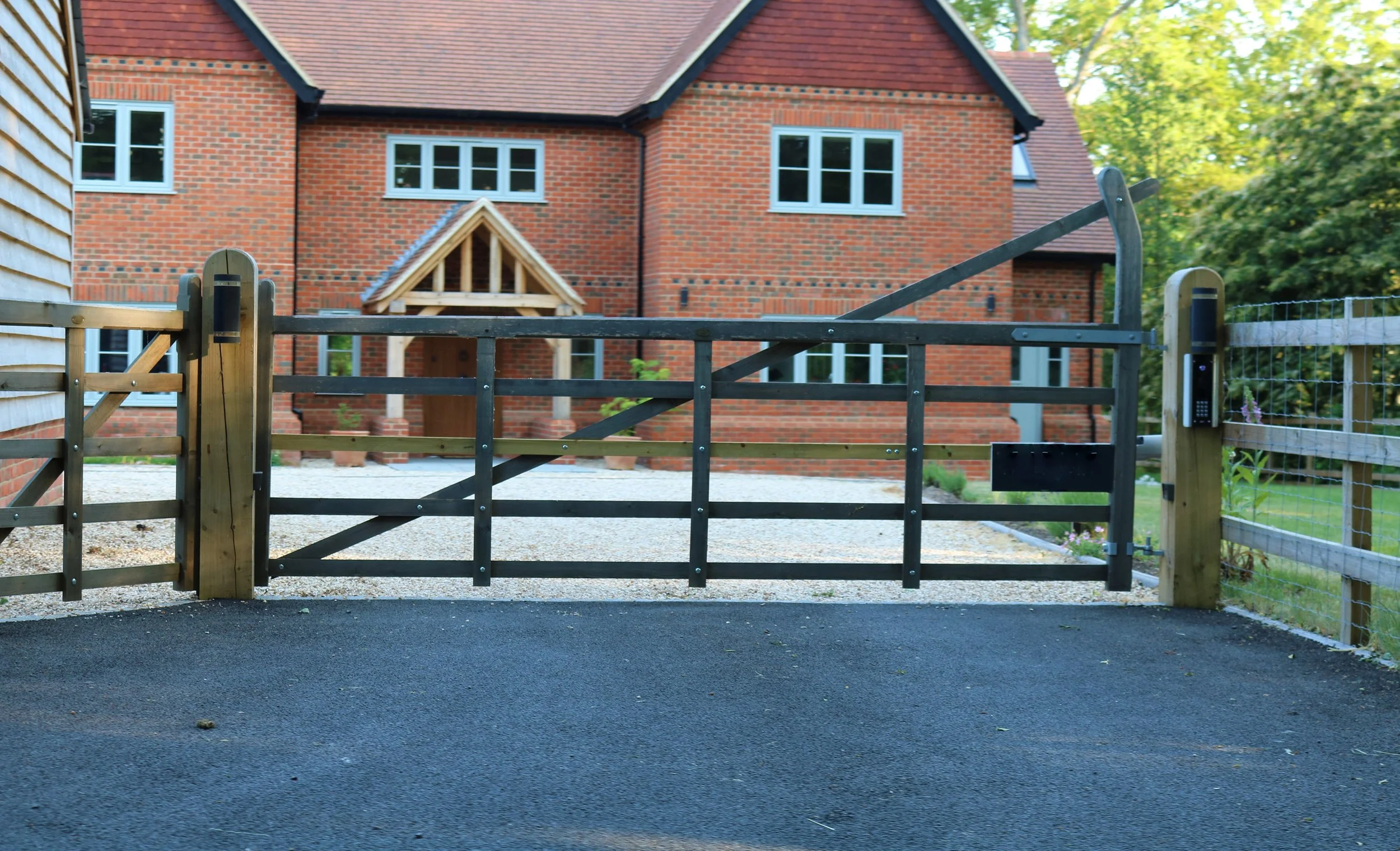 A black driveway gate in front of a new brick house with white windows and a red shingled roof. Surrounding greenery and trees are visible.
