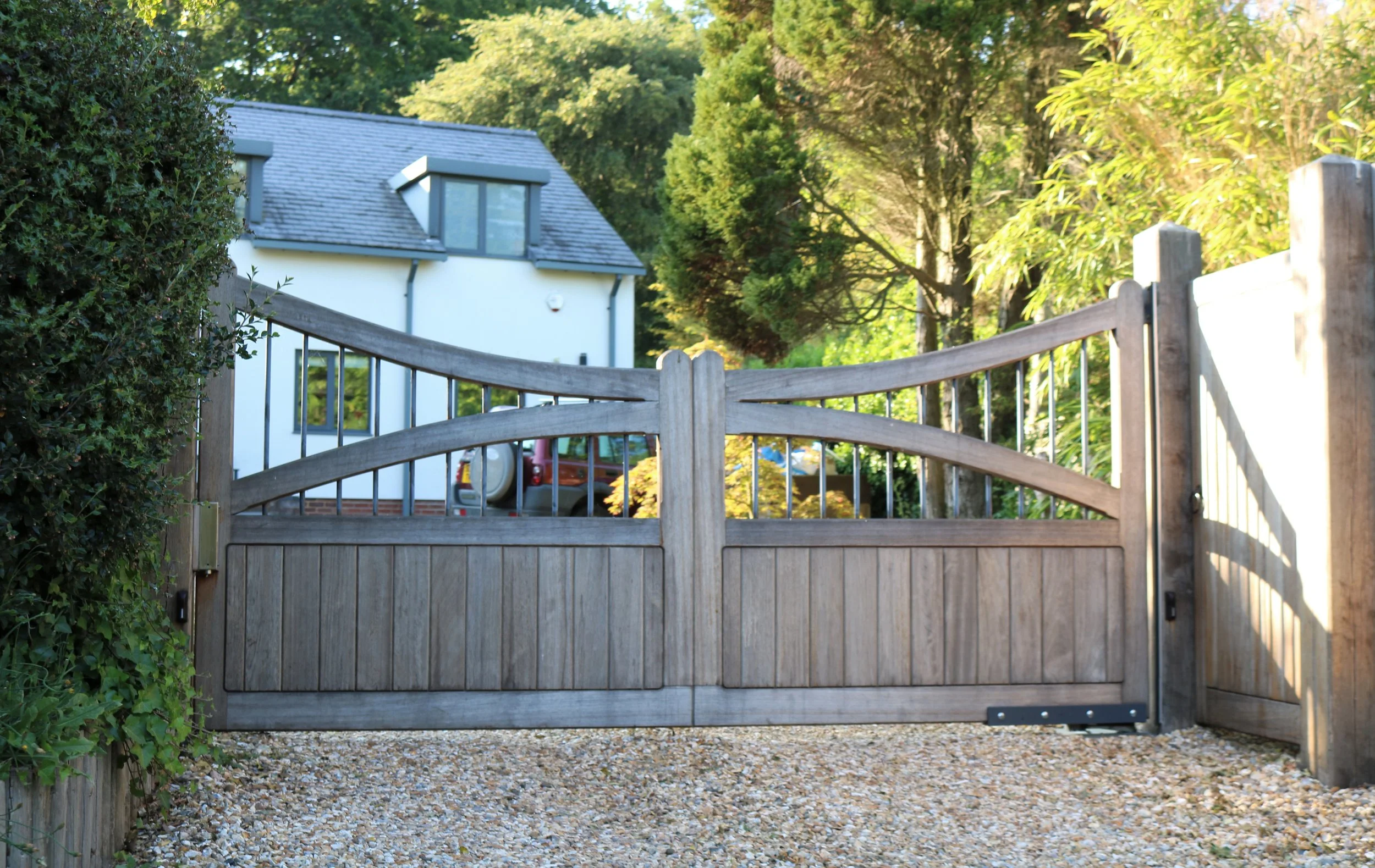 Wooden garden gate with metal hinges, set against a background of a white house with a gray roof, surrounded by green trees and shrubs, with a gravel pathway in front.