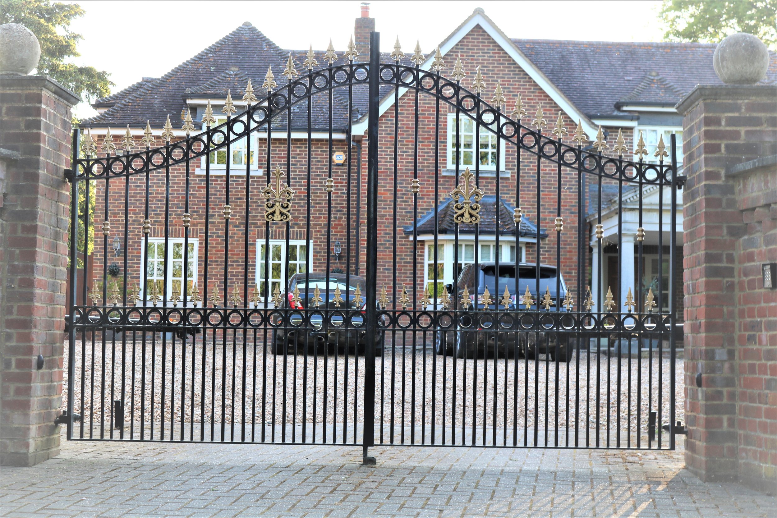 A black wrought iron gate with gold decorative elements, flanked by brick pillars, in front of a brick house with white-trimmed windows and a driveway with cars parked.