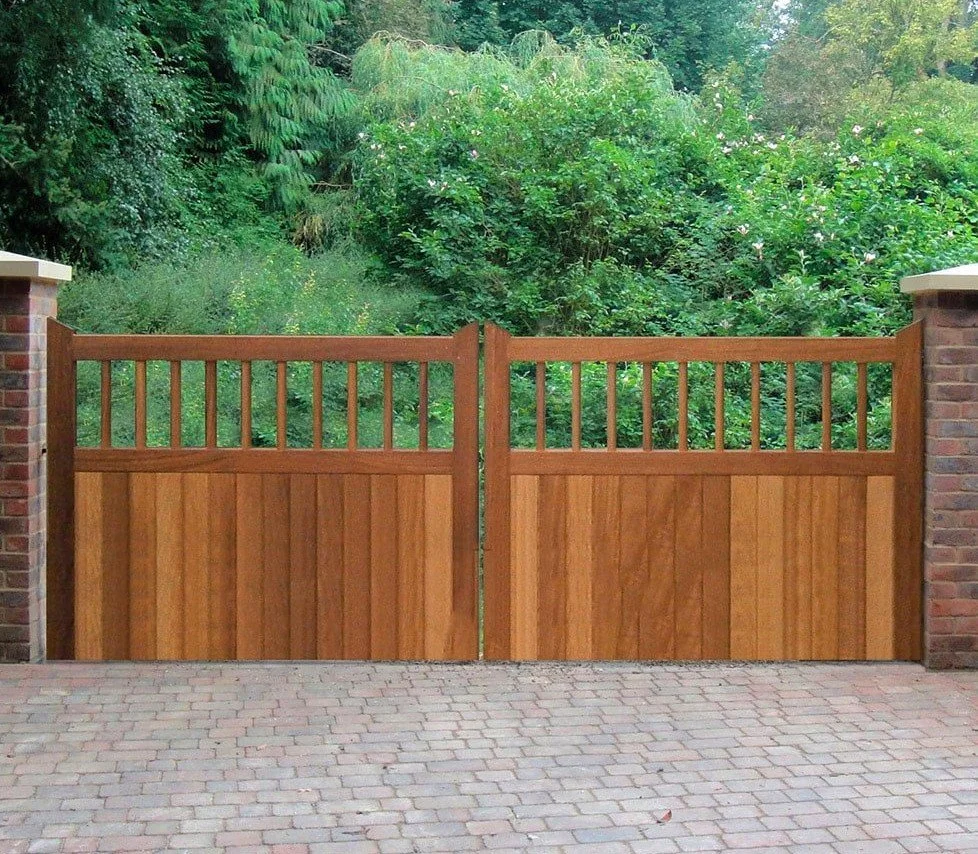 A double wooden gate with vertical slats on the bottom and a horizontal railing at the top, flanked by brick pillars, with greenery and bushes behind it and a brick-paved driveway in front.