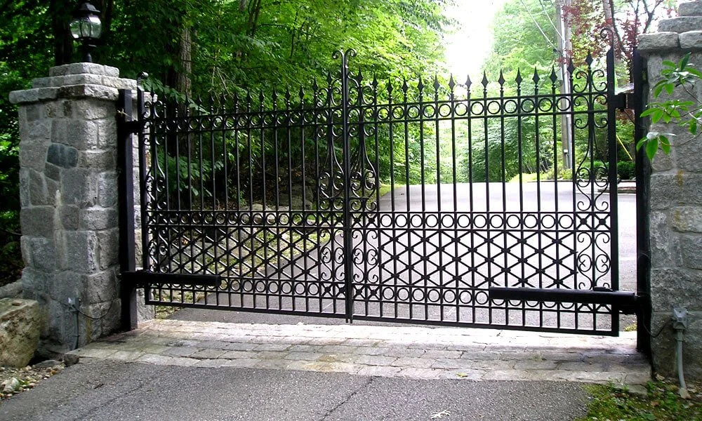 Black wrought iron gate with decorative scrollwork and spear points, set between stone pillars, opening to a paved driveway surrounded by greenery.
