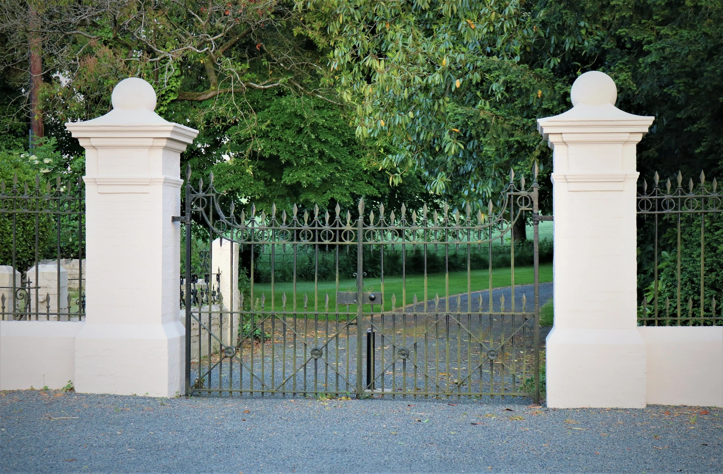 A black wrought iron gate flanked by two white concrete pillars with spherical tops, leading to a lush green garden with tall trees and grass.
