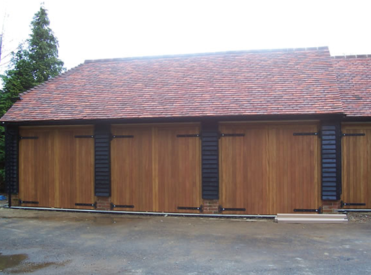 Front view of a wooden garage with black metal hinges and handles, red tile roof, and a gravel driveway in front.