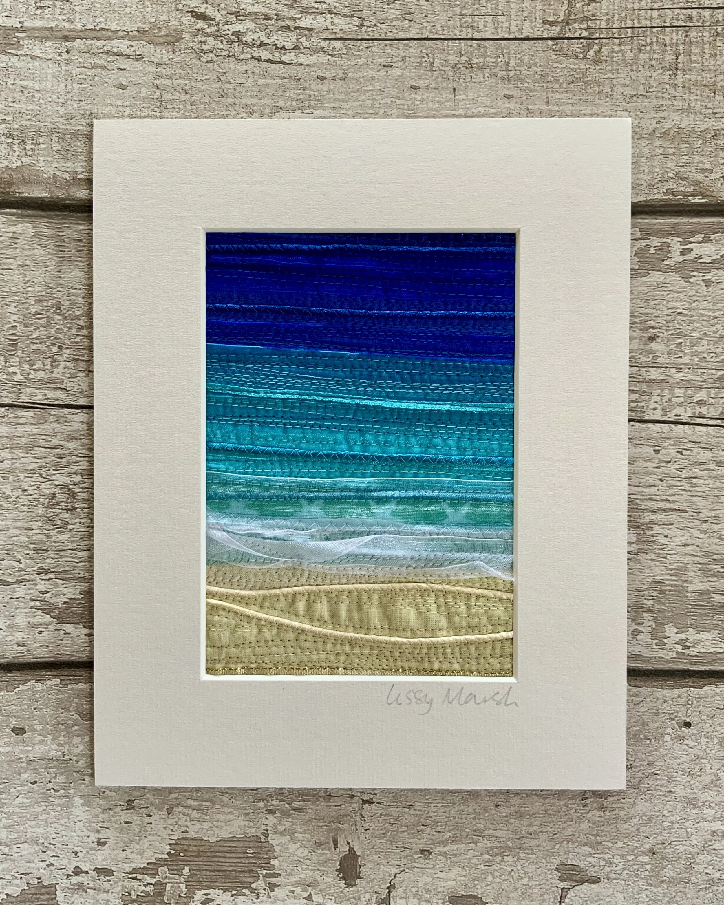 Textile art with layers of blue, white, and beige representing ocean waves and sand, displayed on wooden surface.