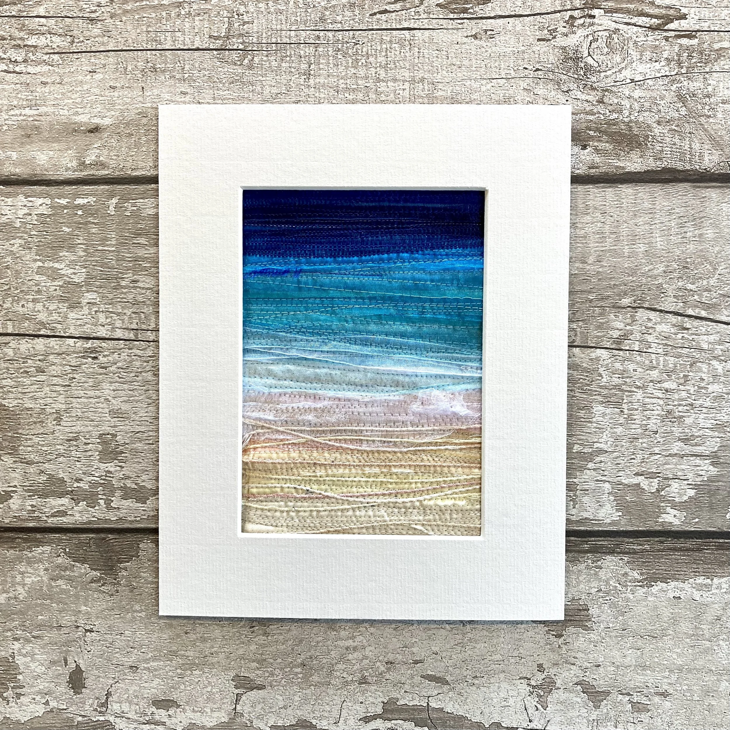Textile art with gradient blue and beige layers, framed and placed on a weathered wooden surface.