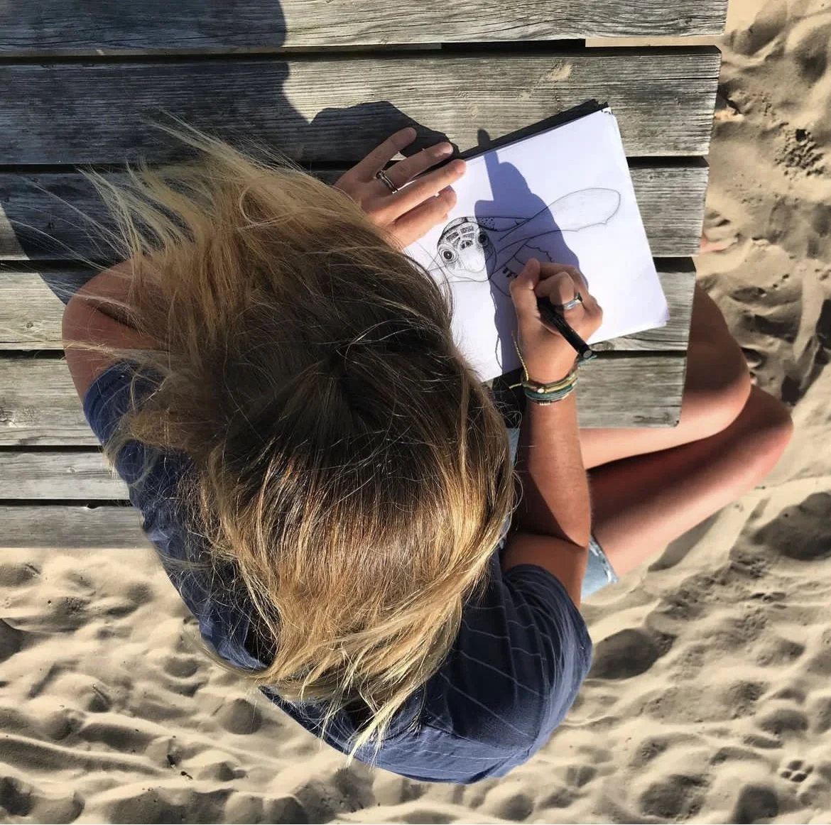 Lissy Marsh drawing on an open sketchbook while sitting on a wooden bench at the beach.