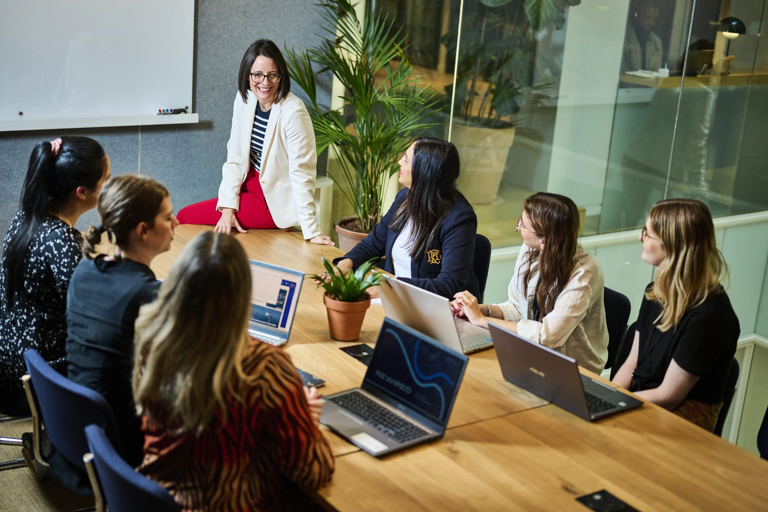 A woman standing at the head of a business meeting table, speaking to a group of six women seated with laptops in front of them. The room has large windows and green plants.
