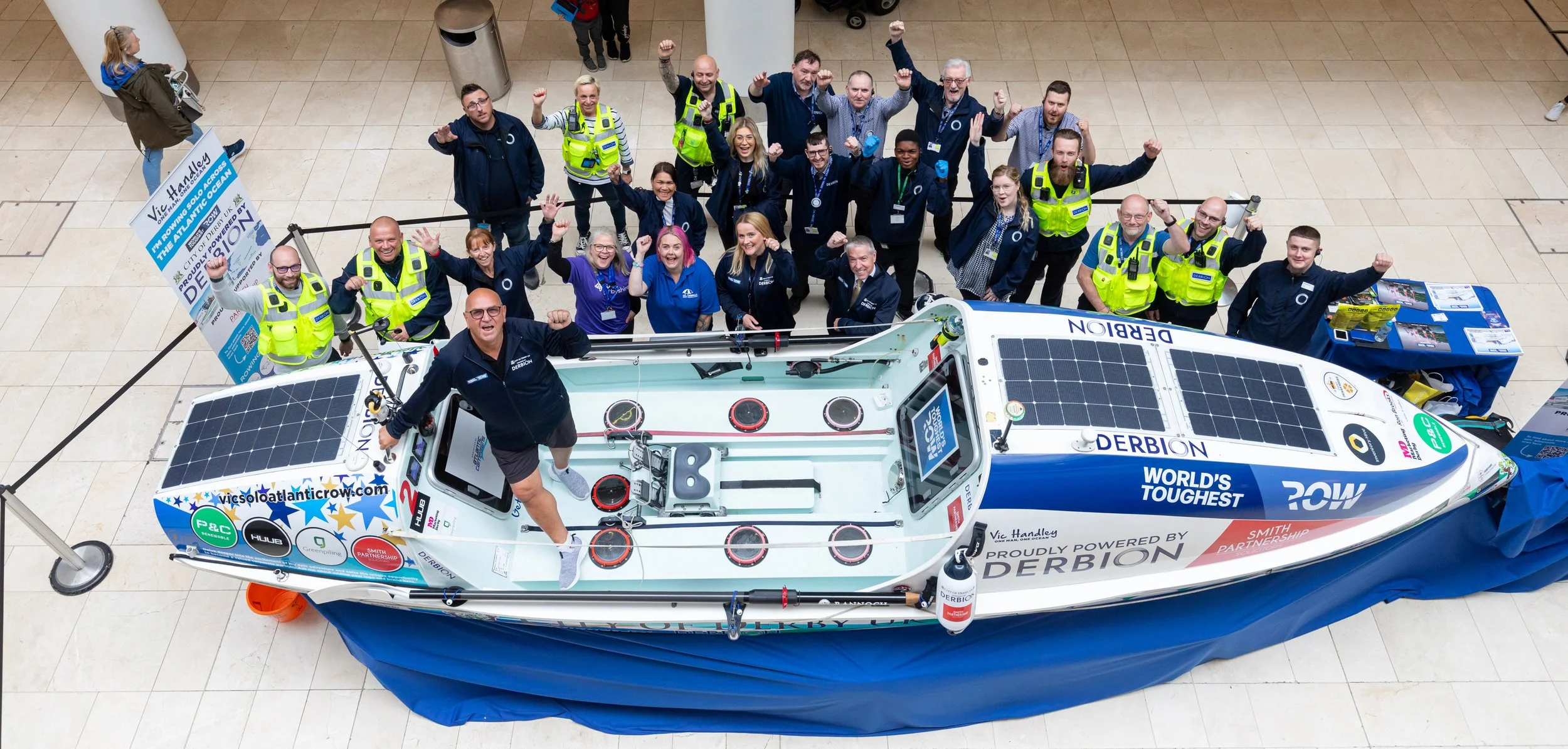 A group of people gathered around a solar-powered boat on display indoors. The team members are smiling and raising their fists in celebration, with some wearing high-visibility vests. The boat features solar panels on its roof and sides, and branding from the Derbion shopping center and various sponsors.