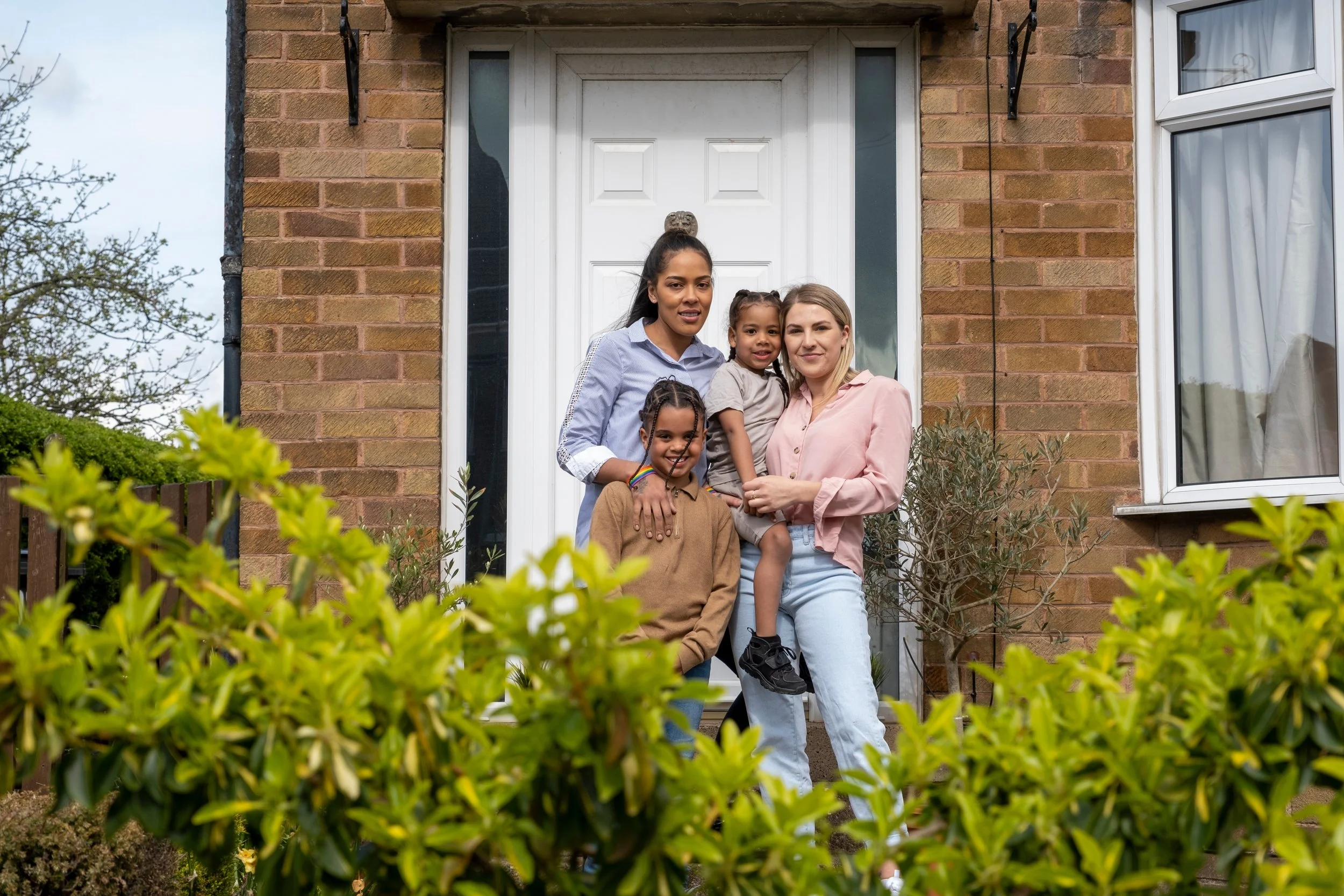 Four women and children stand on a porch in front of a brick house, smiling at the camera, with greenery in the foreground.