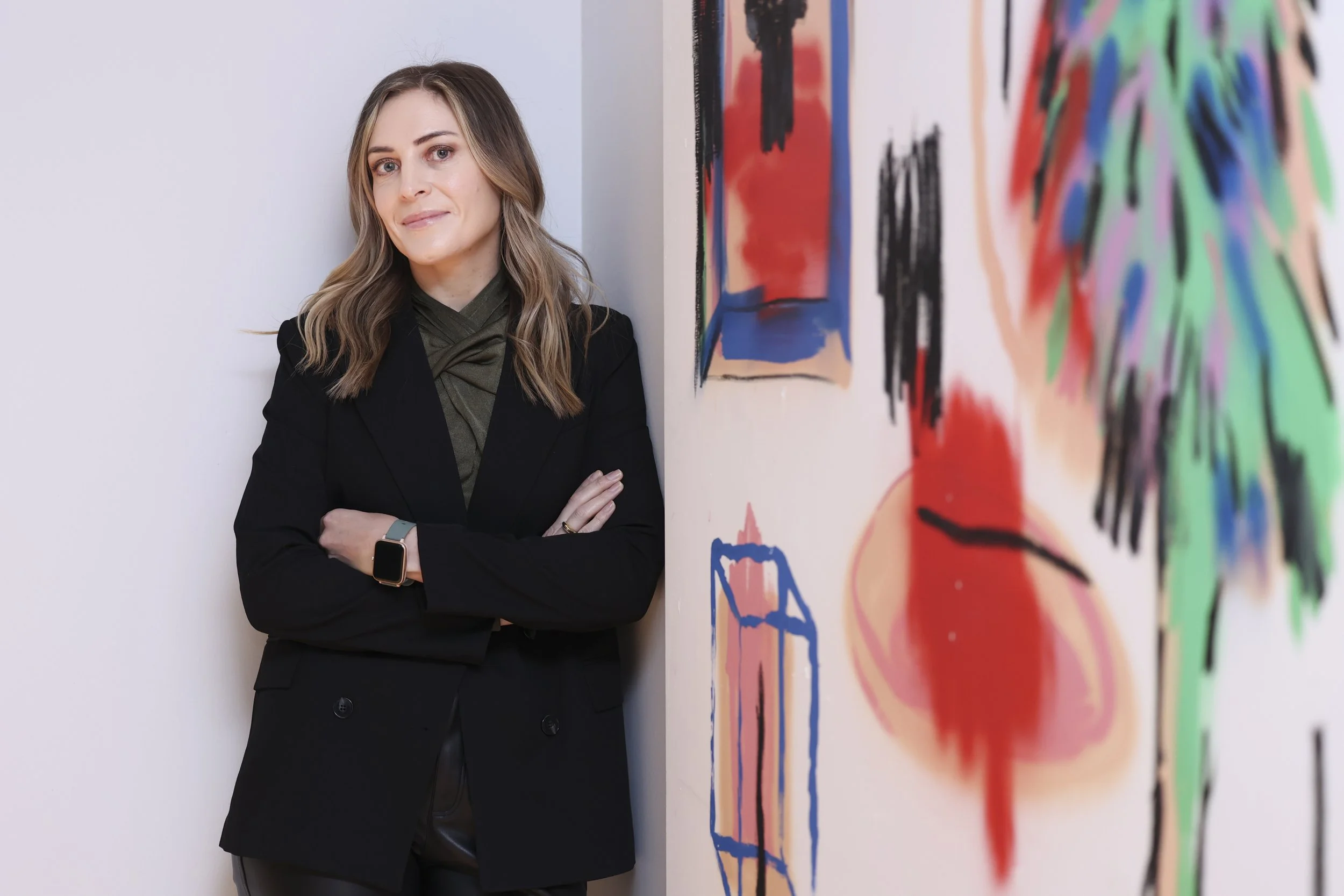 Person with blonde hair, wearing a black blazer, standing next to a wall with abstract colorful artwork in red, blue, and green.