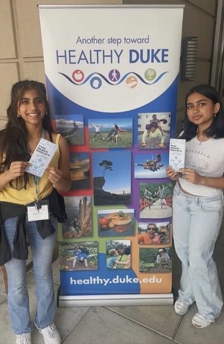 Rhea Kothur and Aanandi Thakur stand on either side of an a standing banner that reads "Another step toward Healthy Duke." They each hold a "Your Health Matters" flyer.