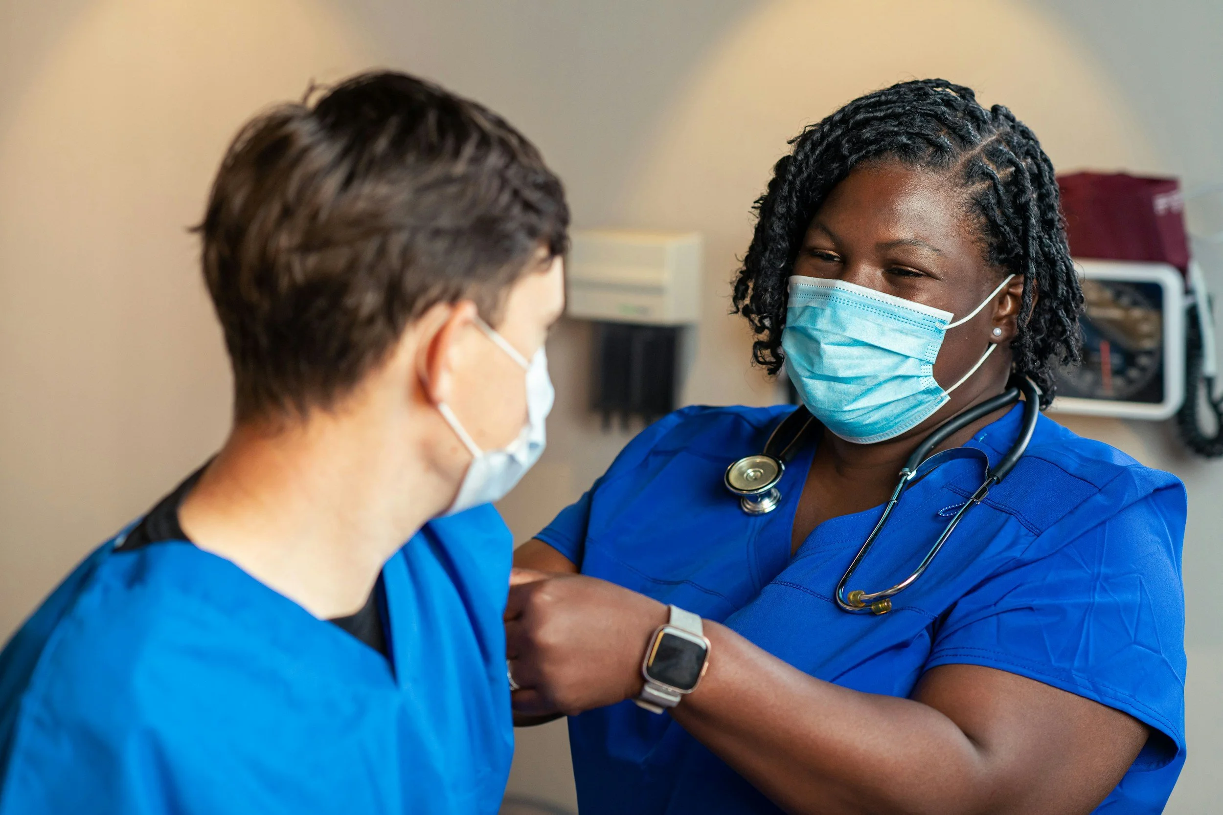 A Black nurse in a dark blue scrubs and a surgical mask attends to a patient in lighter blue, also wearing a surgical mask scrubs