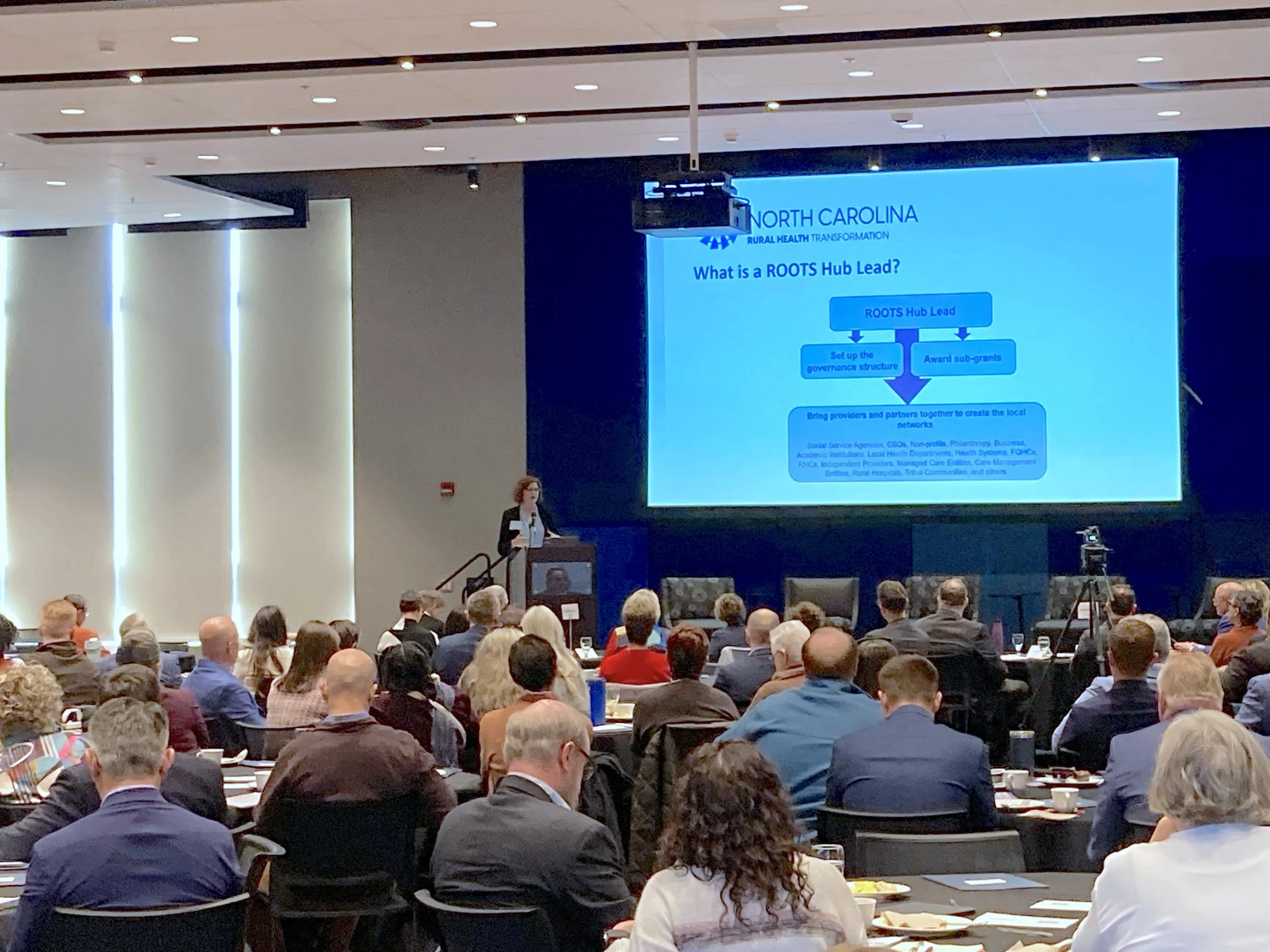 Laurie Stradley (CEO, Impact Health) delivers keynote address “Transforming for Resilience: How Western North Carolina is Reimagining Rural Health.” Showing Roots Hub slide. Photo credit: Michaela Lumpert, Impact Health. 