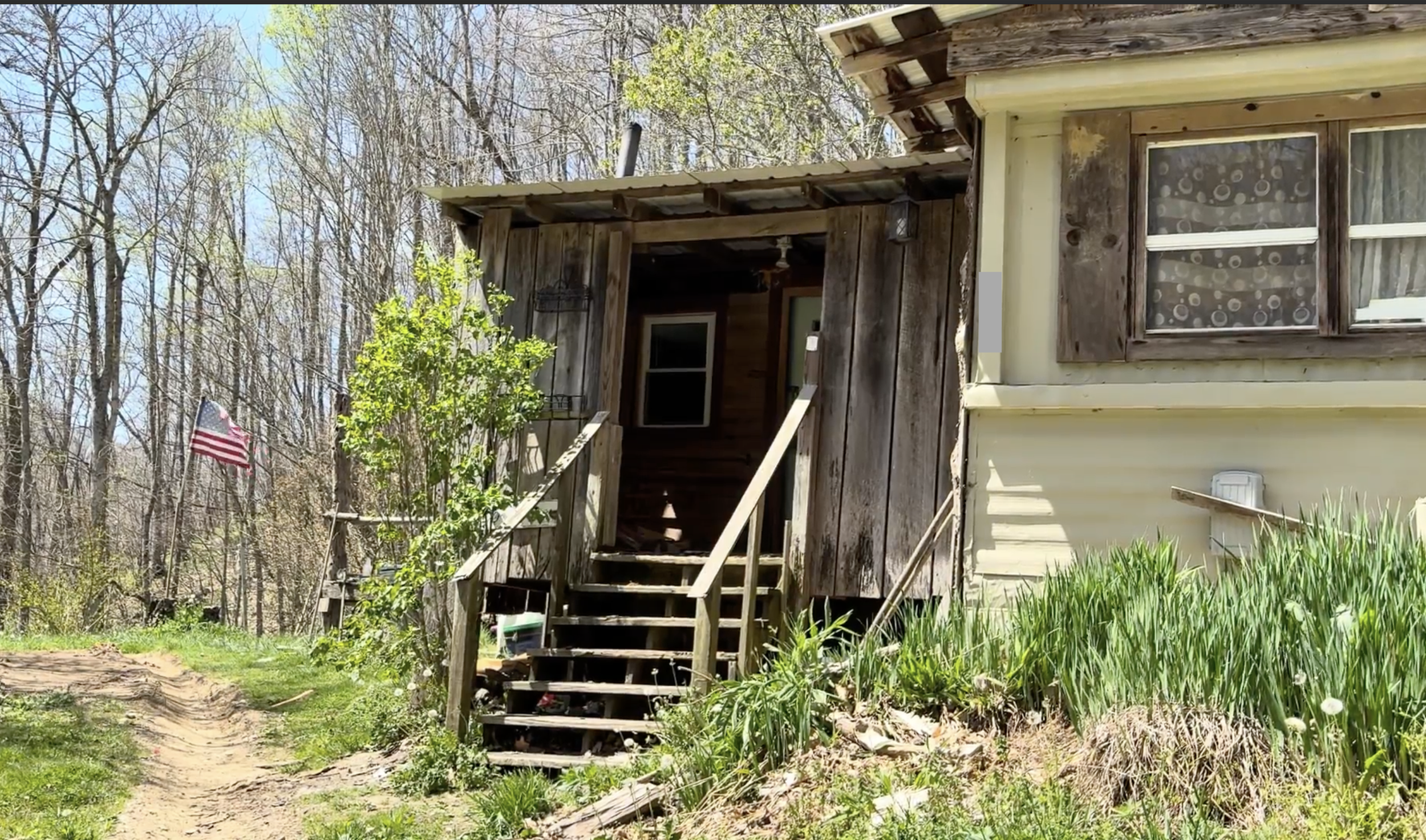 A pale yellow home with an unpainted slat-wood extension sits on wooded mountain side with a dirt path running in front of it.  A pile of dirt overgrown with grass is mounded in front of the home. An American flag flies on a pole in the background.