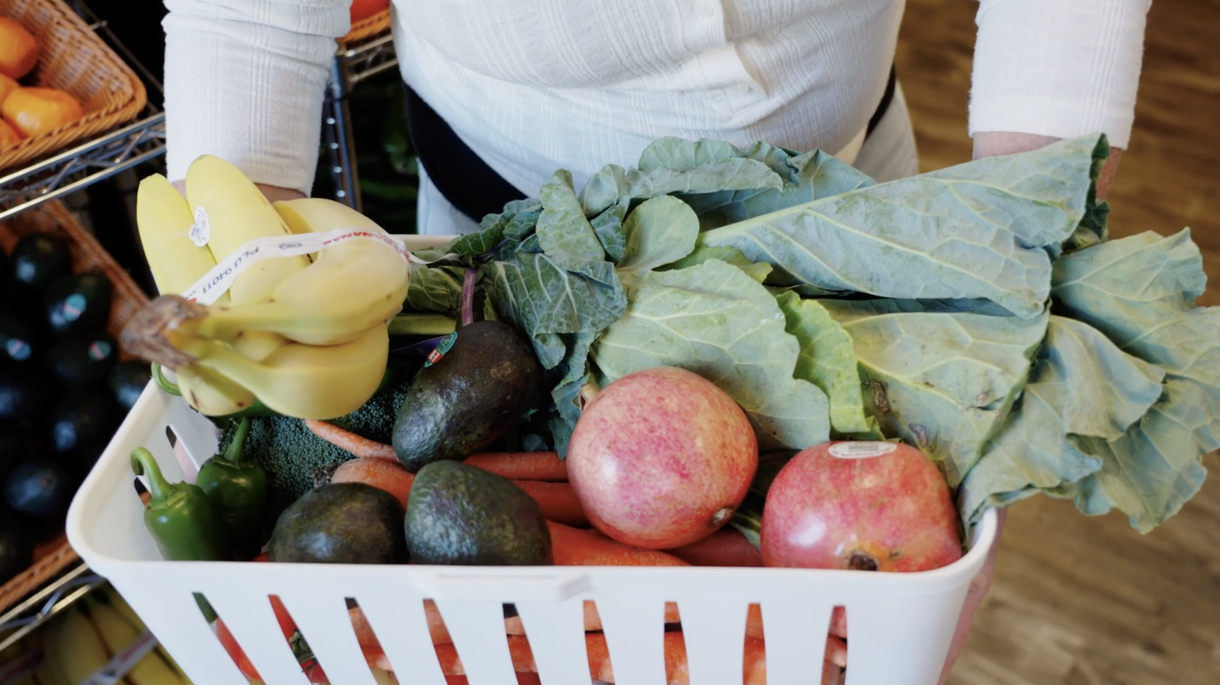 A person in a white shirt holds a white basket full of fresh fruits and vegetables. Shelves containing baskets of produce can be seen in the background.