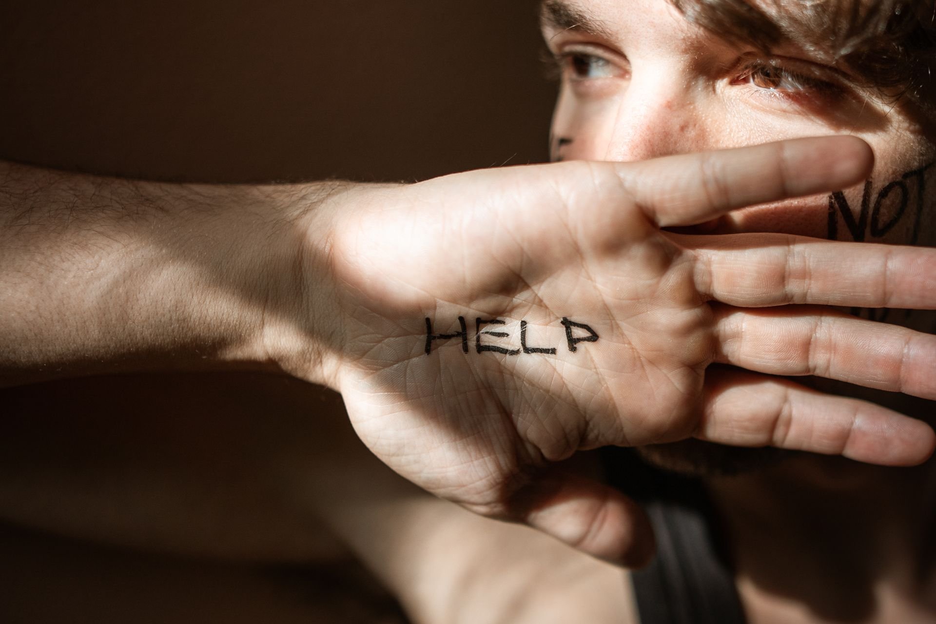 Close-up of a person holding their hand in front of their face, palm facing out. The word "HELP" is written on their palm in black ink.