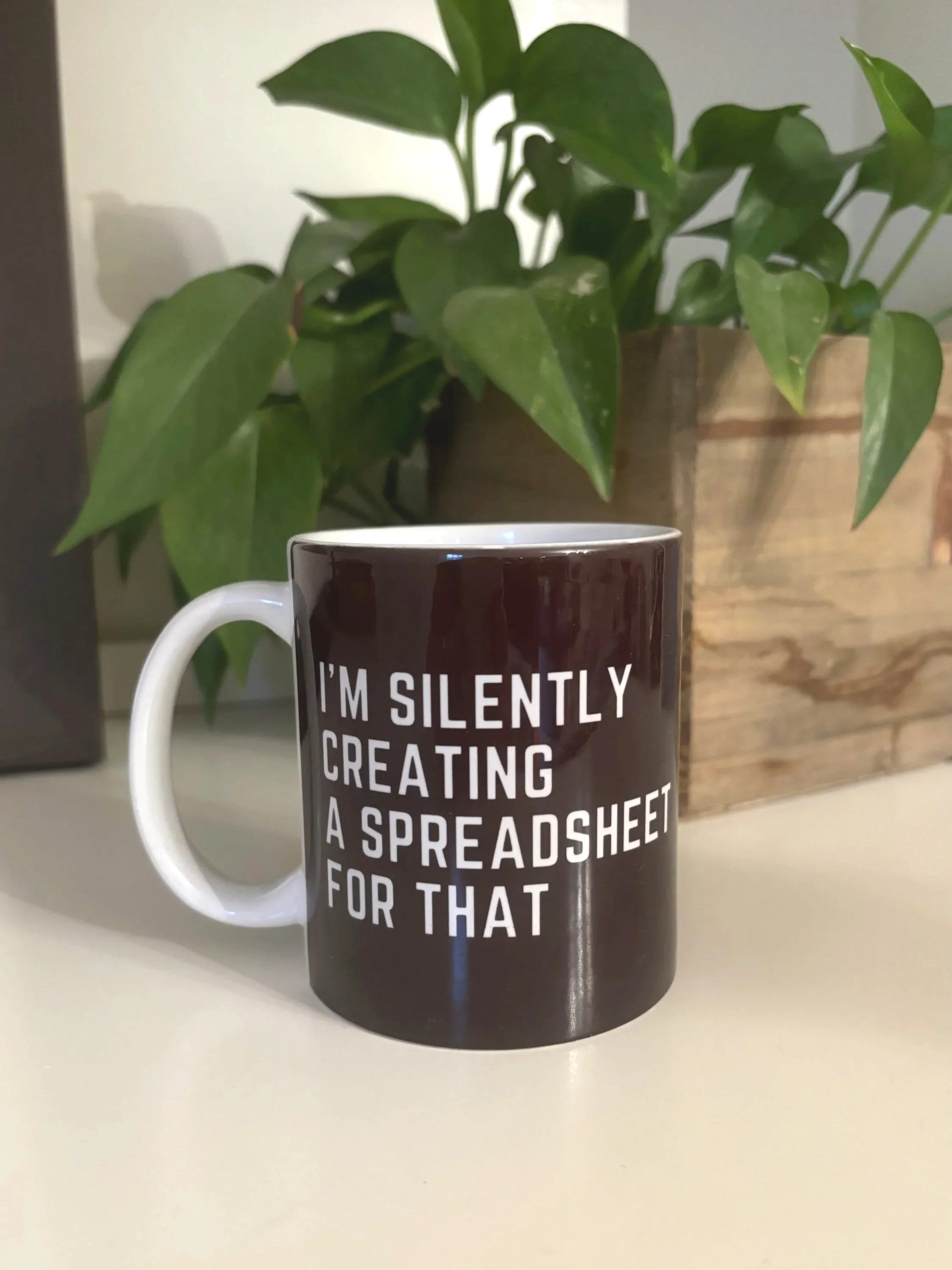 Black coffee mug with the phrase “I’m silently creating a spreadsheet for that,” sitting on a desk next to a plant.