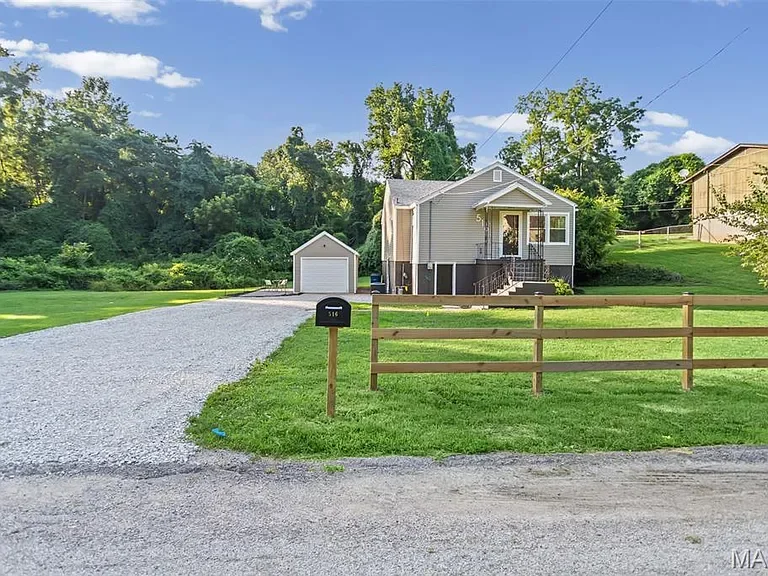 A suburban house with a gravel driveway, green lawn, wooden fence, and detached garage. Full exterior home remodel with new roof and vinyl siding in Alton Illinois renovated by ASA Construction. 