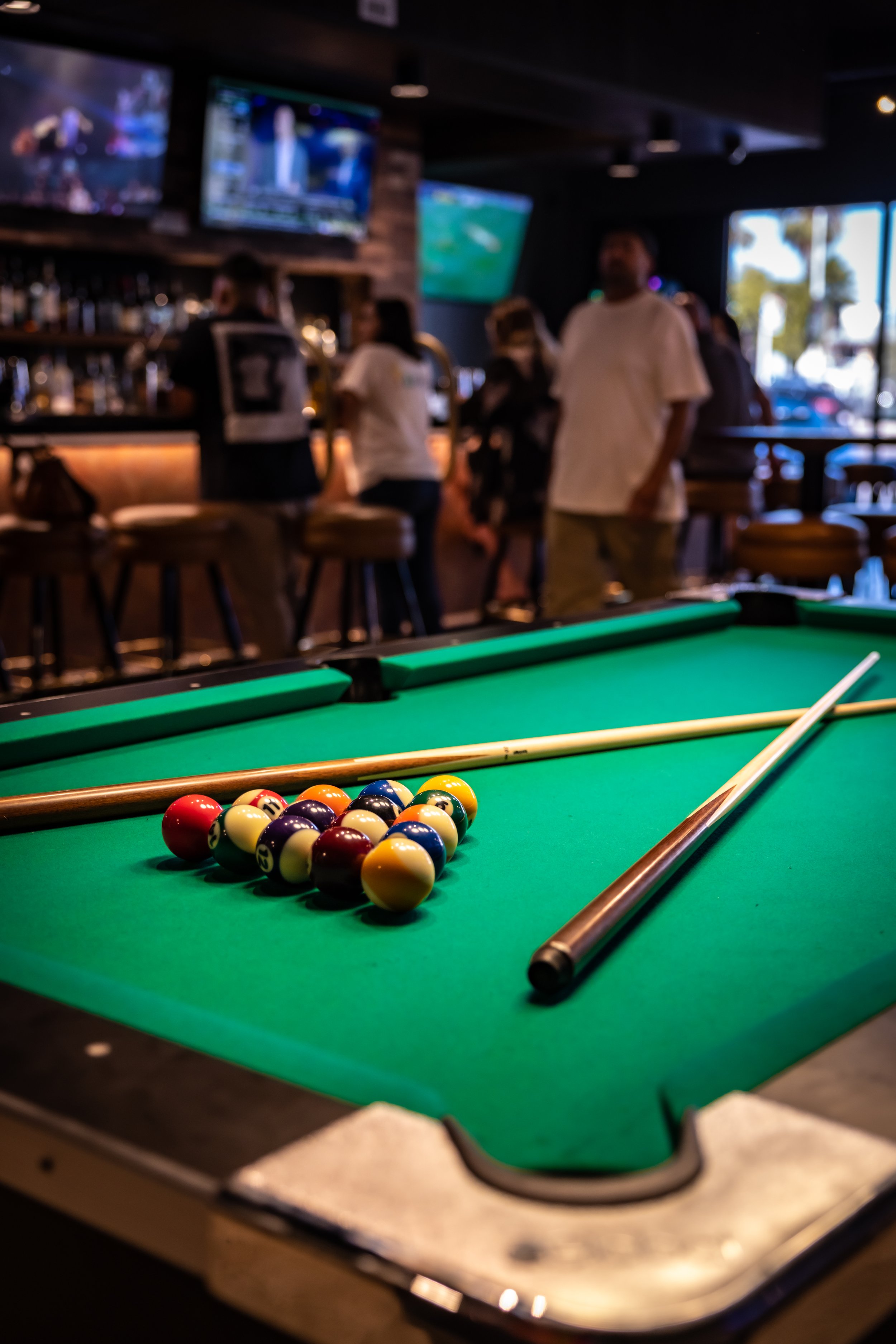 Pool table with billiard balls racked and two cues, inside a bar with people gathered around the bar and television screens in the background.