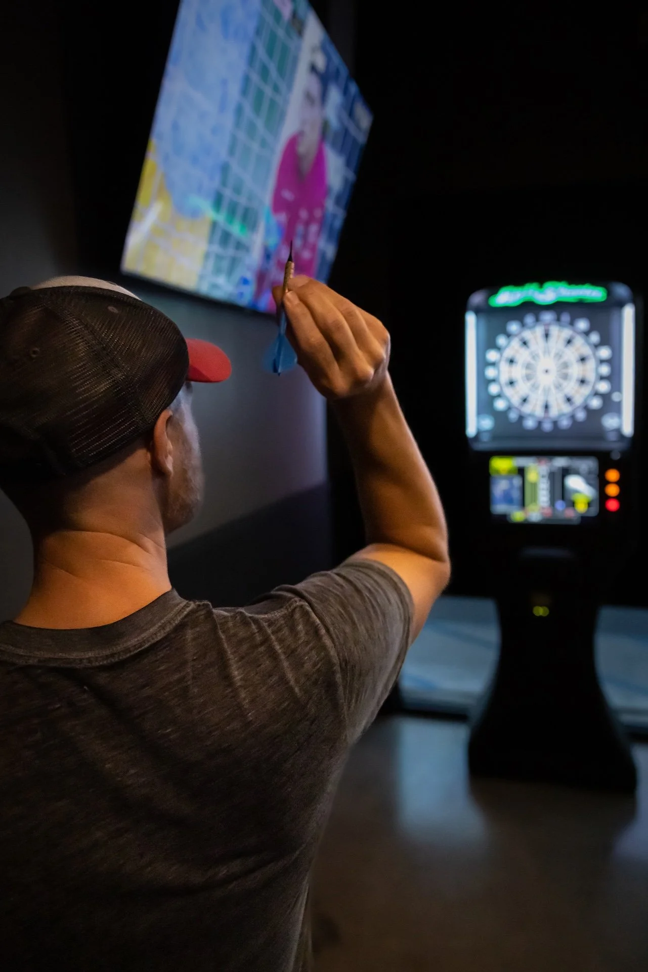 A person wearing a baseball cap, viewed from behind, is throwing a dart at a dartboard in an indoor setting with a dark background. There is a screen displaying a colorful image of a person and an electronic dartboard with lights in the background.