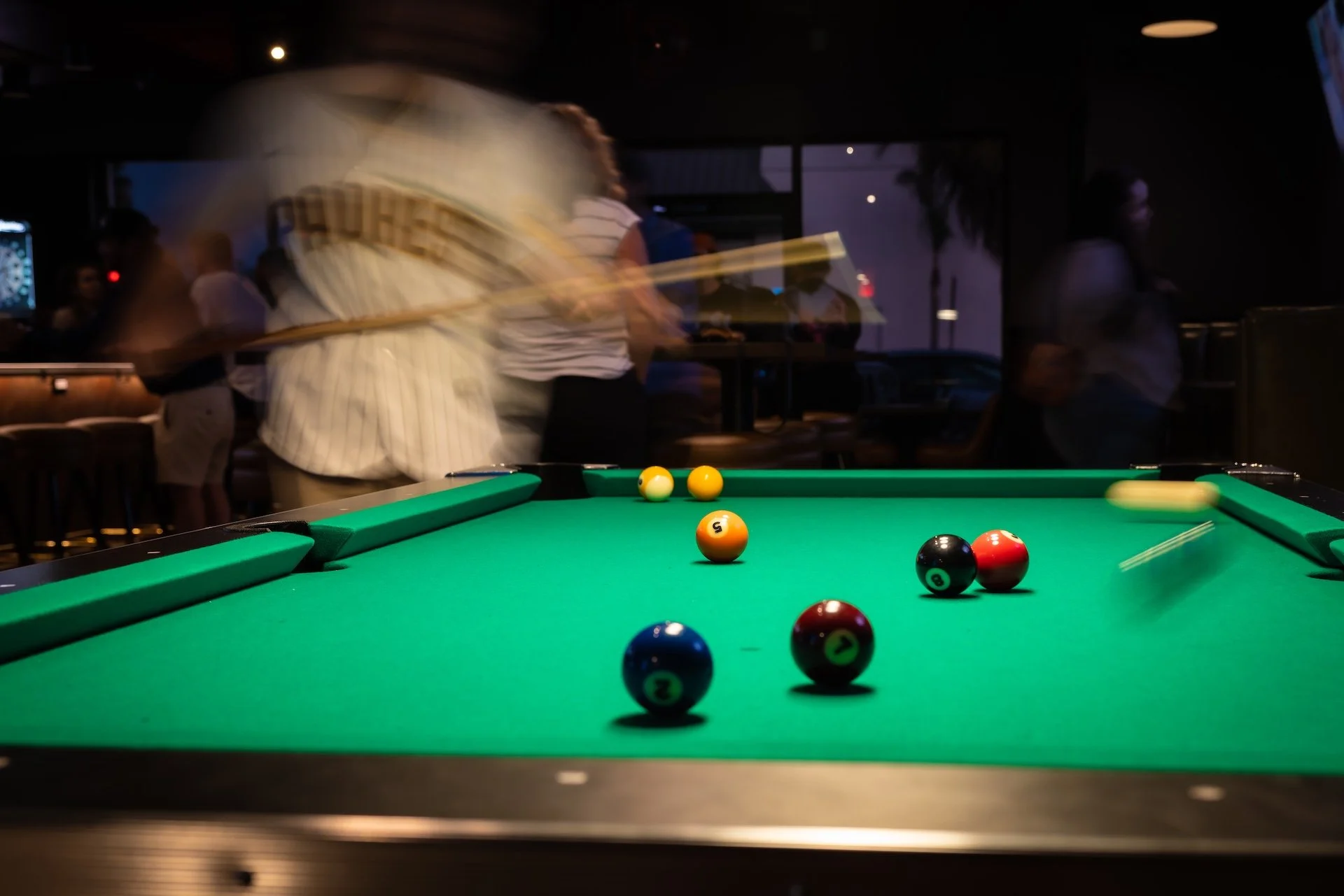 A pool table with billiard balls on it in a dimly lit room with people in the background, some moving quickly, creating a blurred effect.
