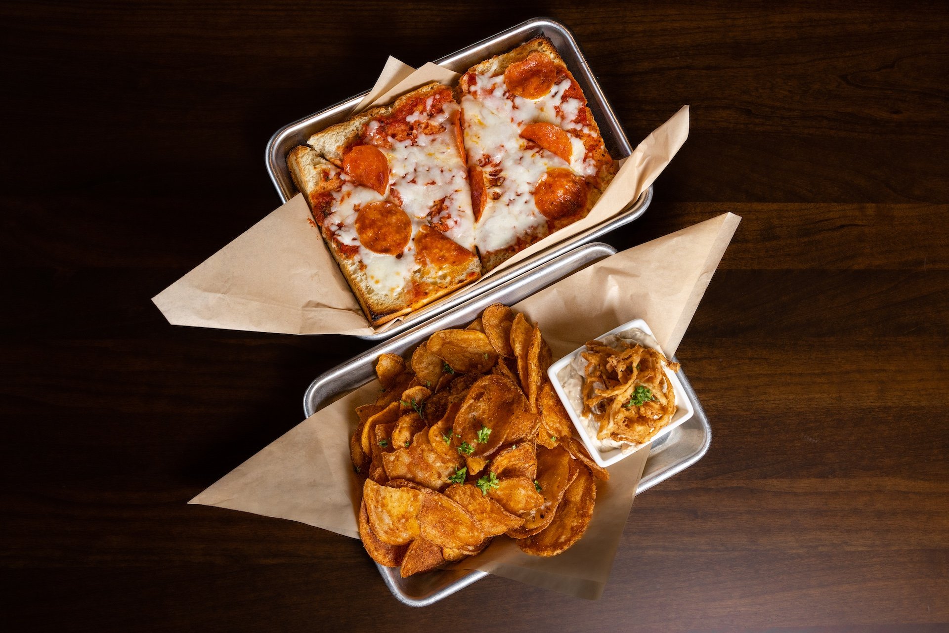 Top view of two metal trays with pub food on a dark wood table. The top tray has a slice of pepperoni pizza with melted cheese. The bottom tray has a serving of potato chips, a small bowl of fried onion strings, and a side of ranch dip.