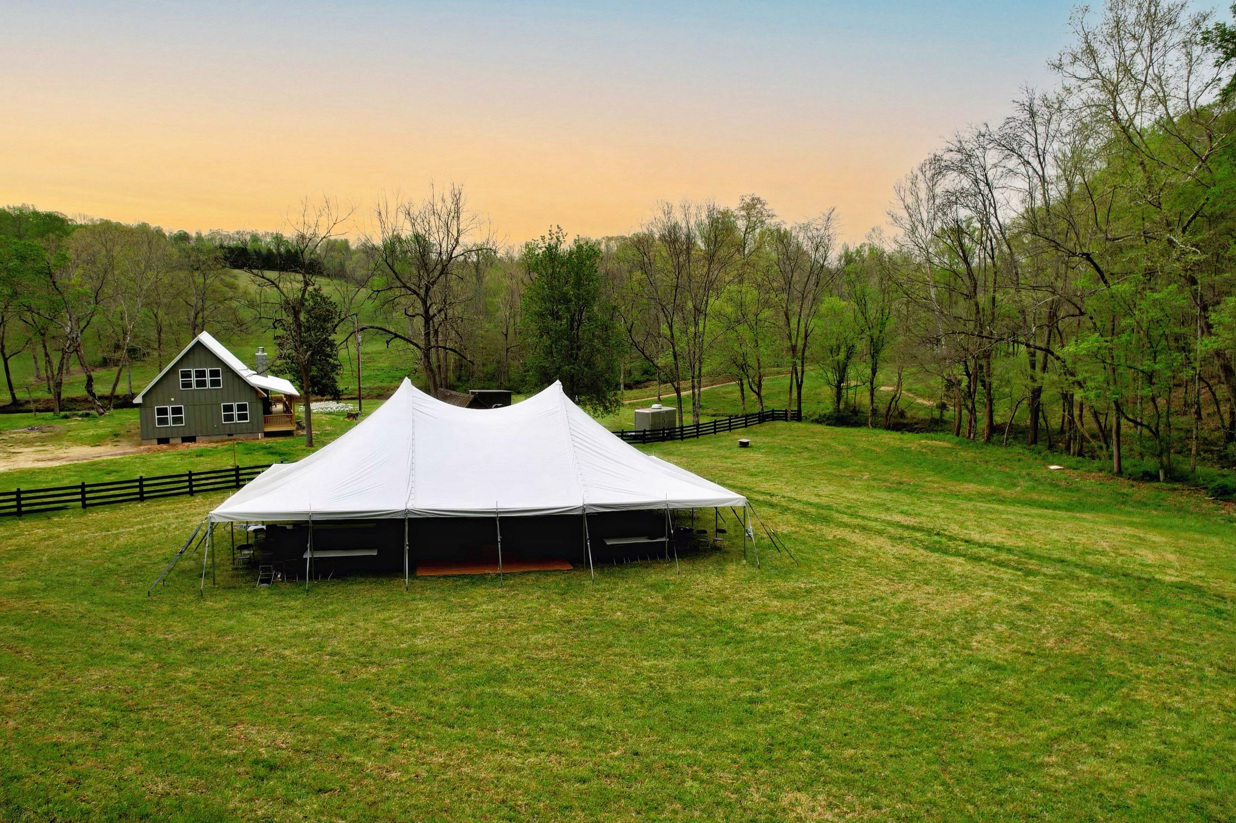Large white event tent set up on a grassy lawn, with a small green house in the background and trees surrounding the area during sunset.