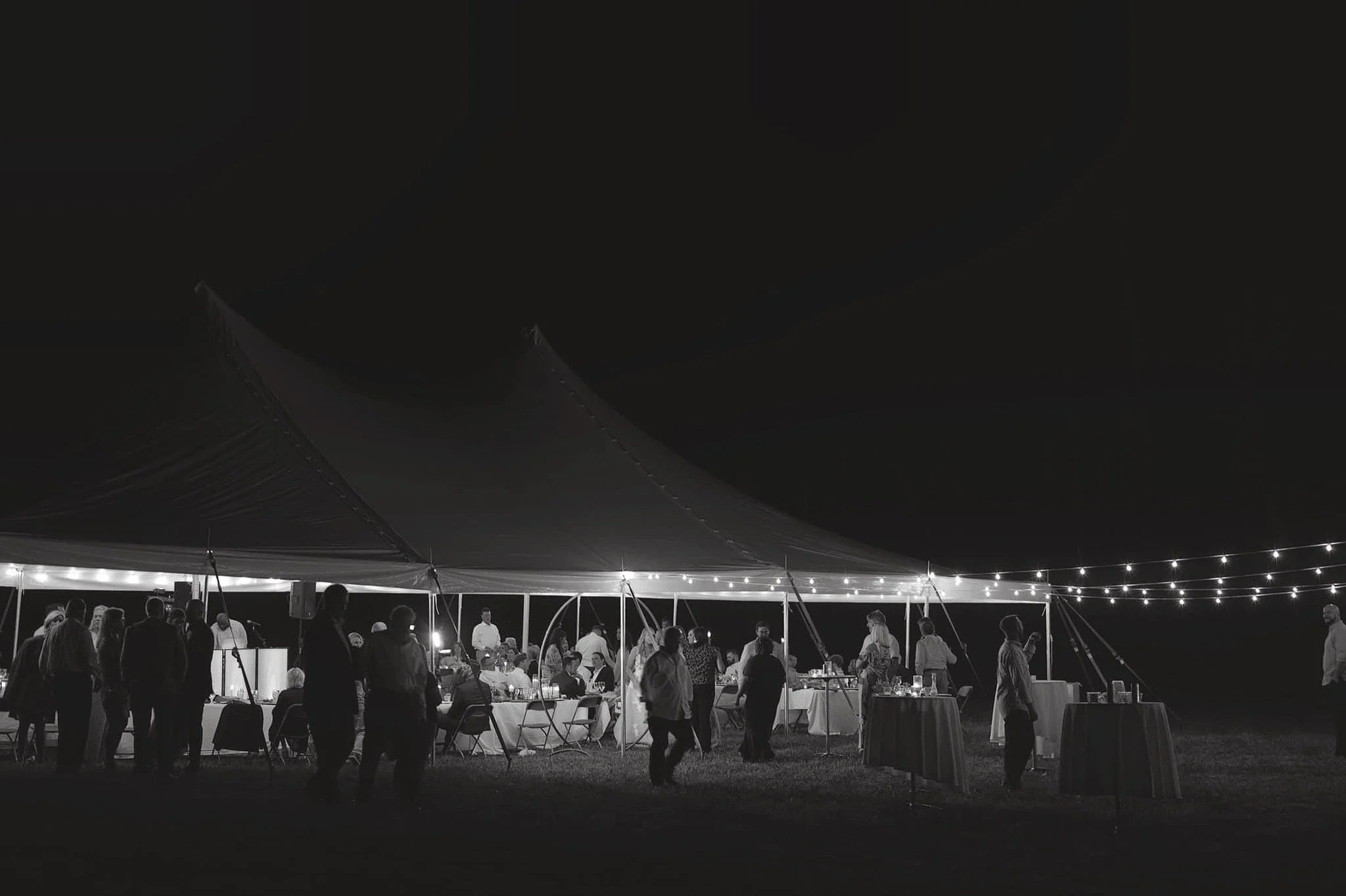 Nighttime outdoor gathering under a large tent with string lights, people socializing and dining at tables.