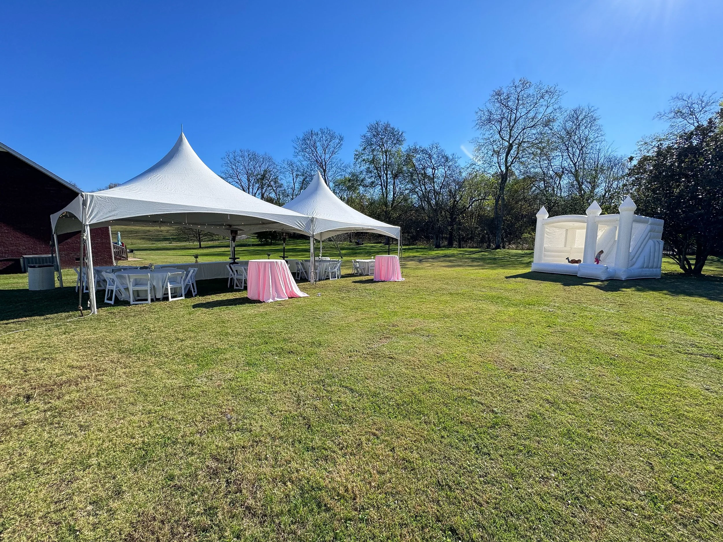 Outdoor event setup with white tents, pink table skirts, and a white inflatable slide on a grassy field with trees in the background on a sunny day.
