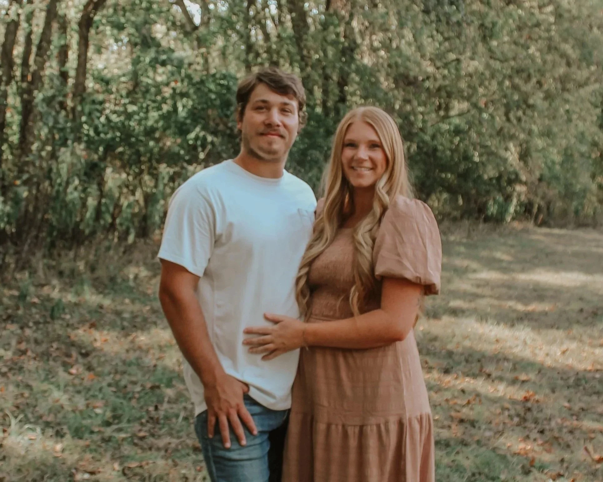 A smiling couple standing in a wooded outdoor area.