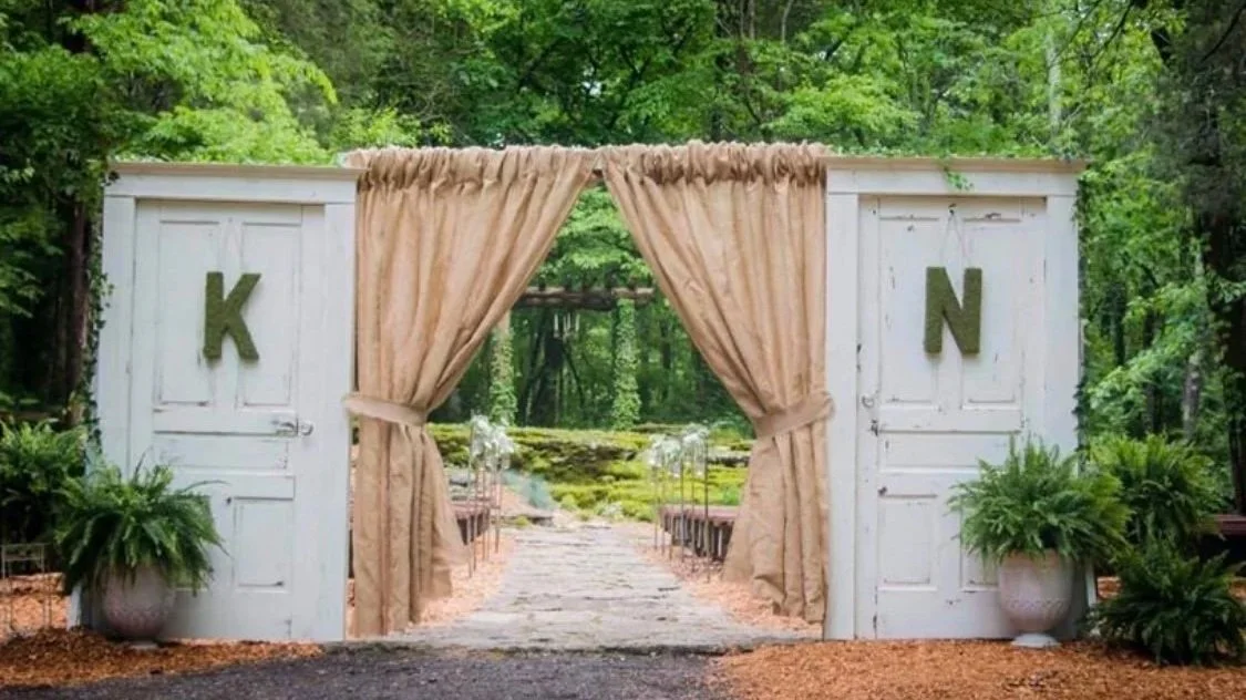 Entrance to an outdoor wedding venue with white wooden doors, beige curtains, and greenery, with the letters K and N on the doors.