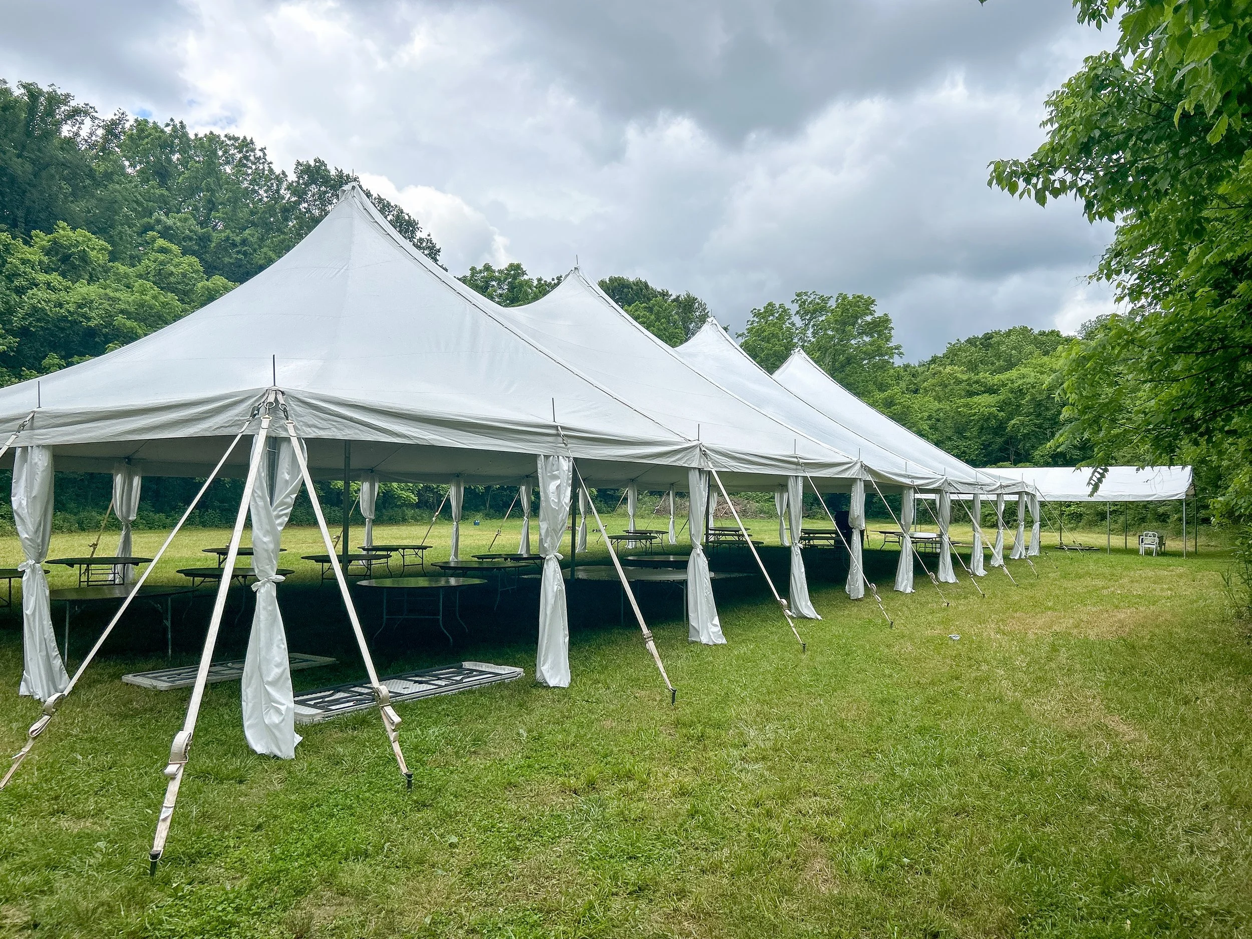 Large outdoor white event tent set up on grassy field with trees and cloudy sky in the background.