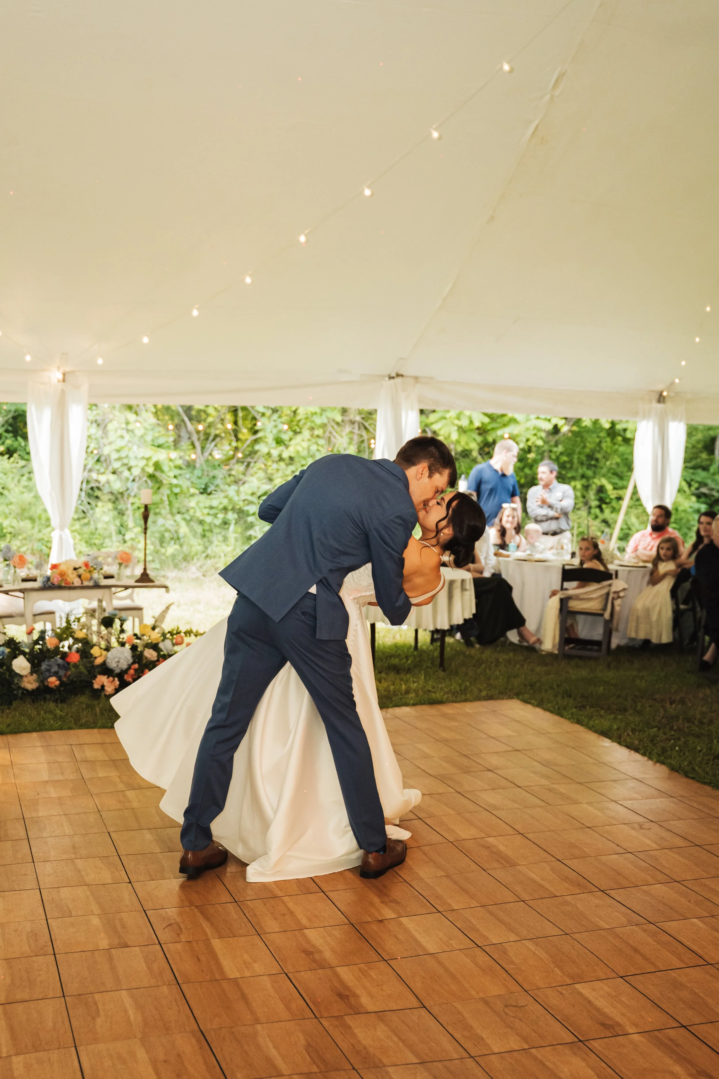 A bride and groom sharing a kiss during their wedding reception under a decorated tent with guests watching.