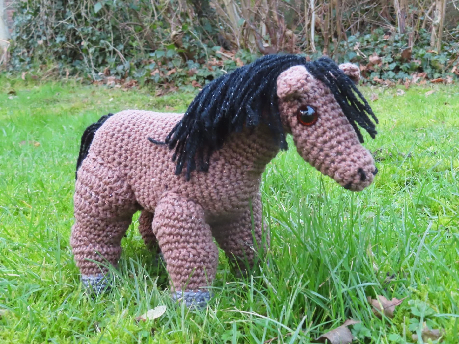 A brown crocheted horse with a black mane on a grassy lawn