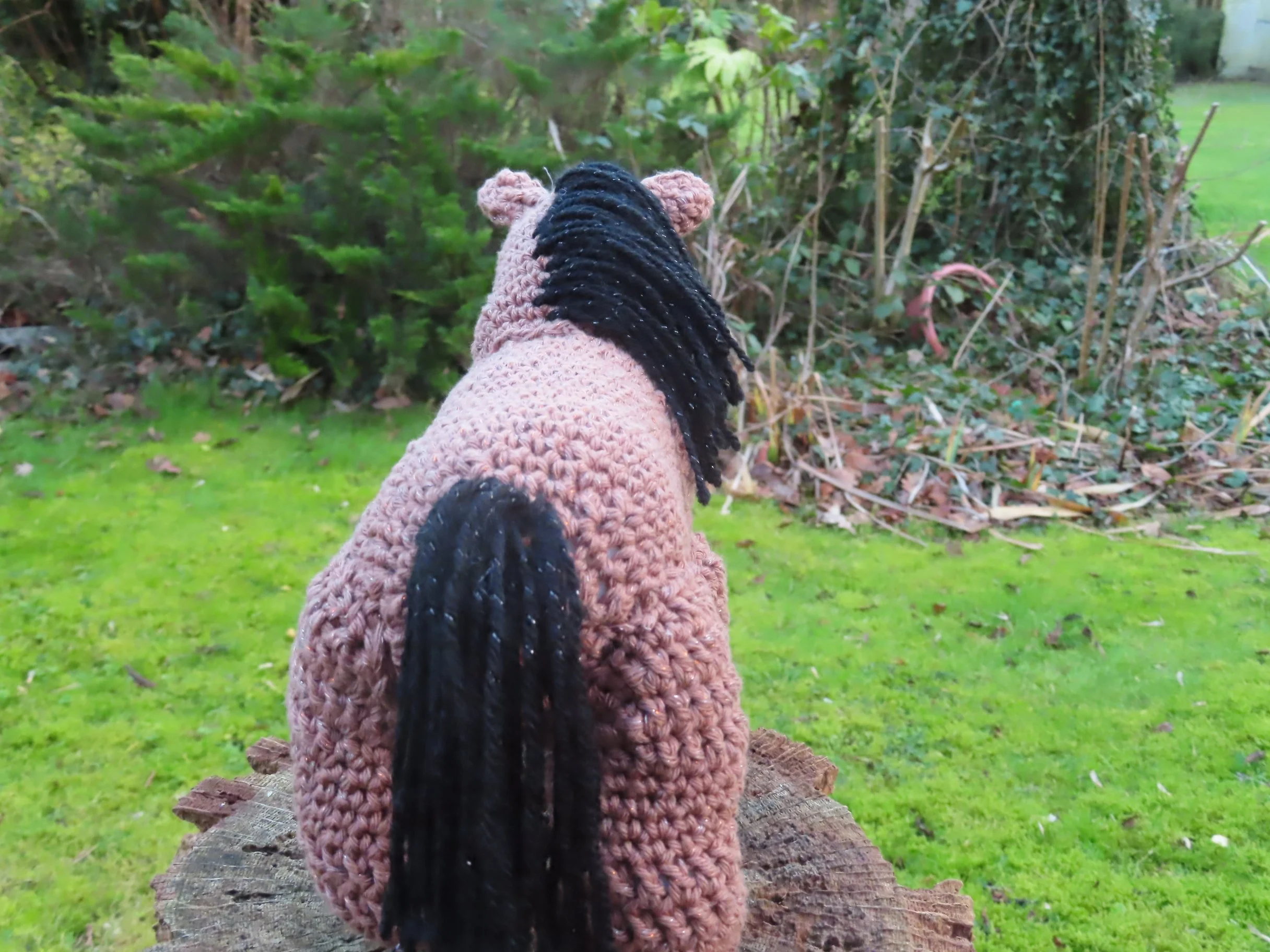 A brown crocheted horse with a black mane looking out on a garden