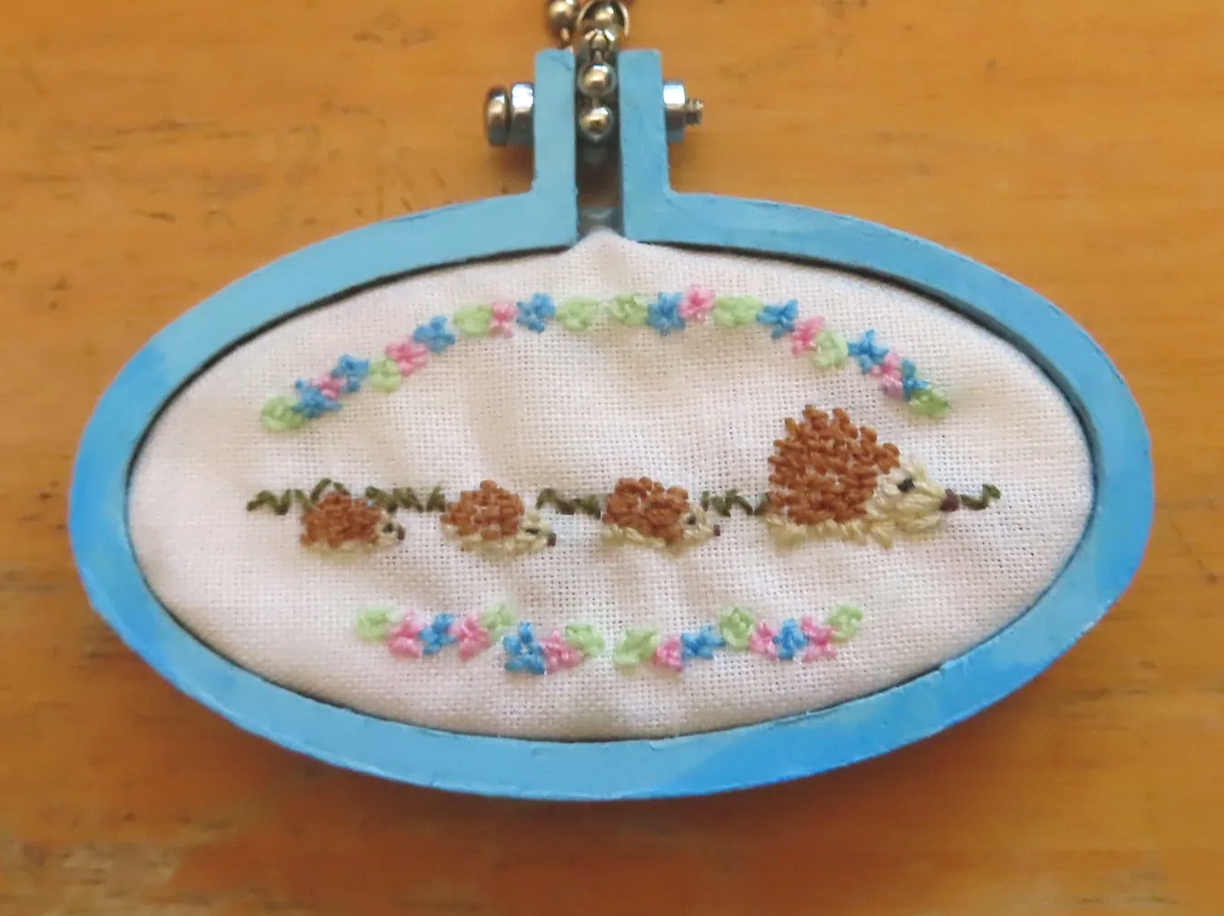 A small piece of embroidery depicting a mother hedgehog and three baby hedgehogs in a line, in a light blue oval frame