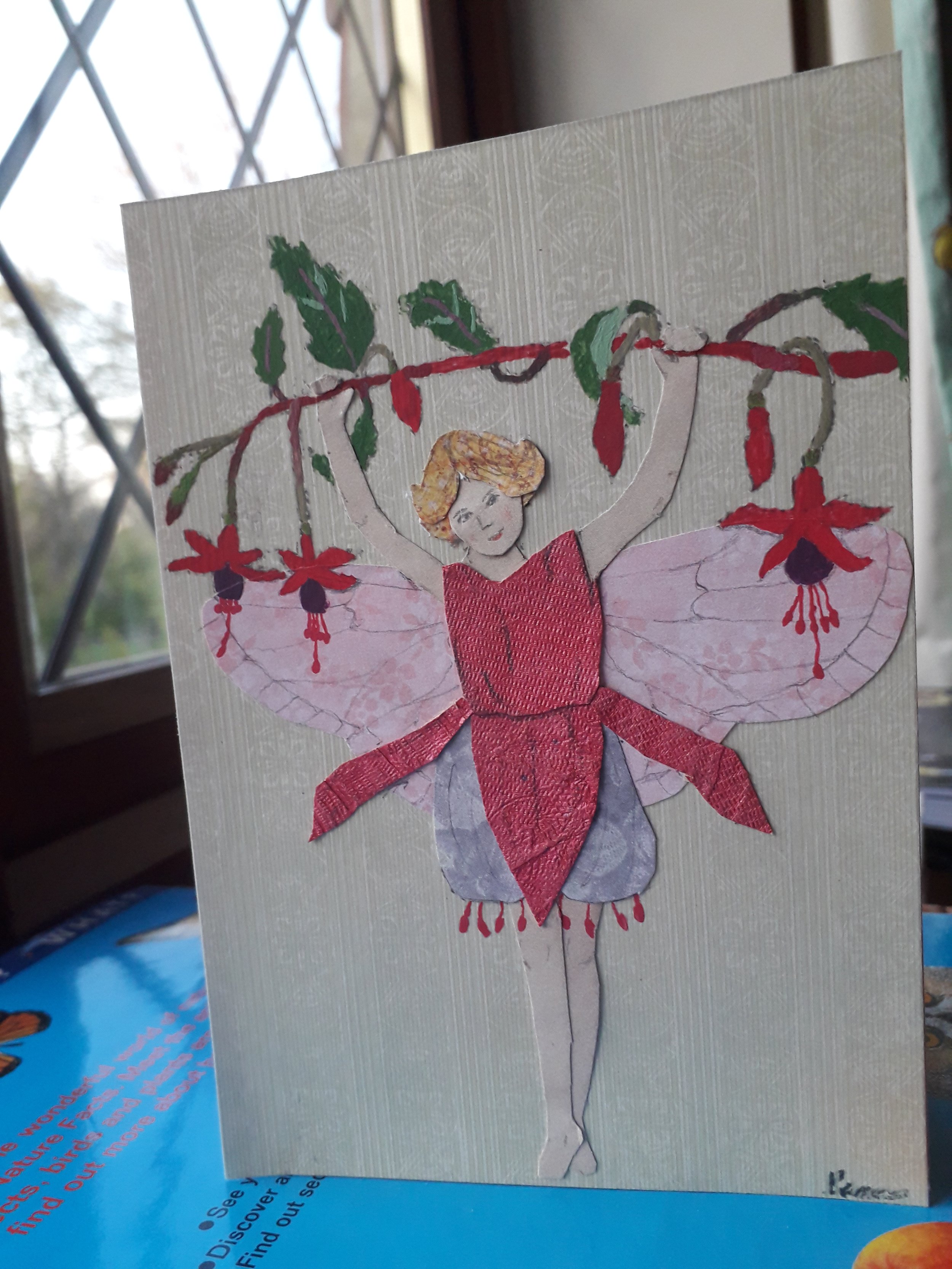 A handmade greetings card with a collage design depicting a fairy with a dress made from a fuchsia flower holding onto a fuchsia stalk