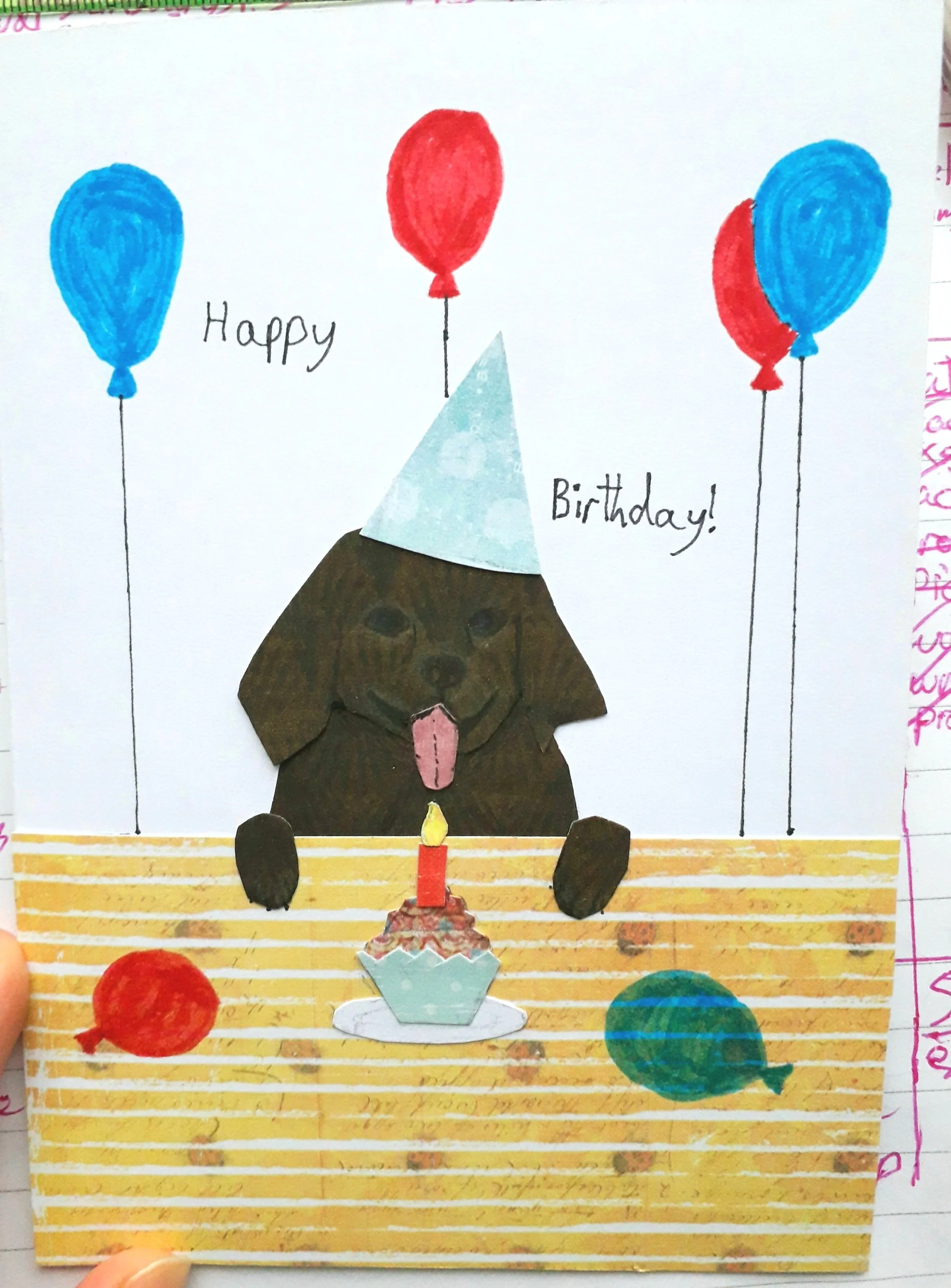 A handmade card depicting a brown Labrador puppy wearing a party hat with a cupcake in front of it, surrounded by balloons, and the words "Happy Birthday!"